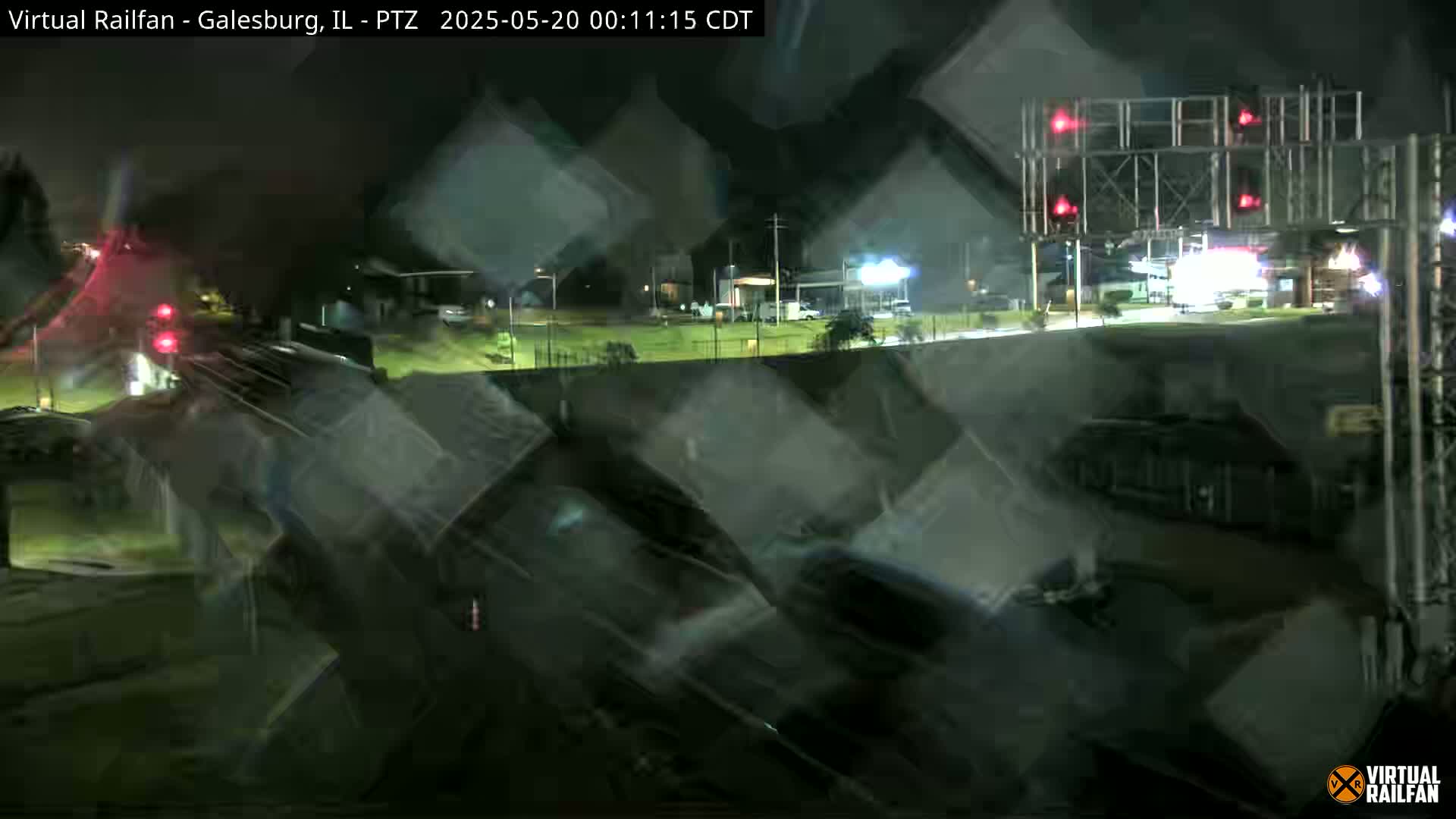 A nighttime view of a train passing under a railroad crossing with flashing red lights, showing vehicles on a road nearby.