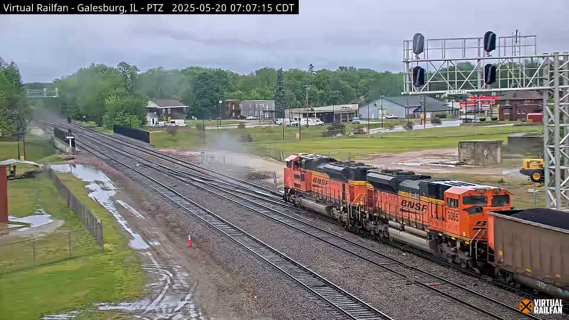 A long orange freight train passes through a flat landscape under an overcast sky, with puddles visible on the ground near the railway tracks.