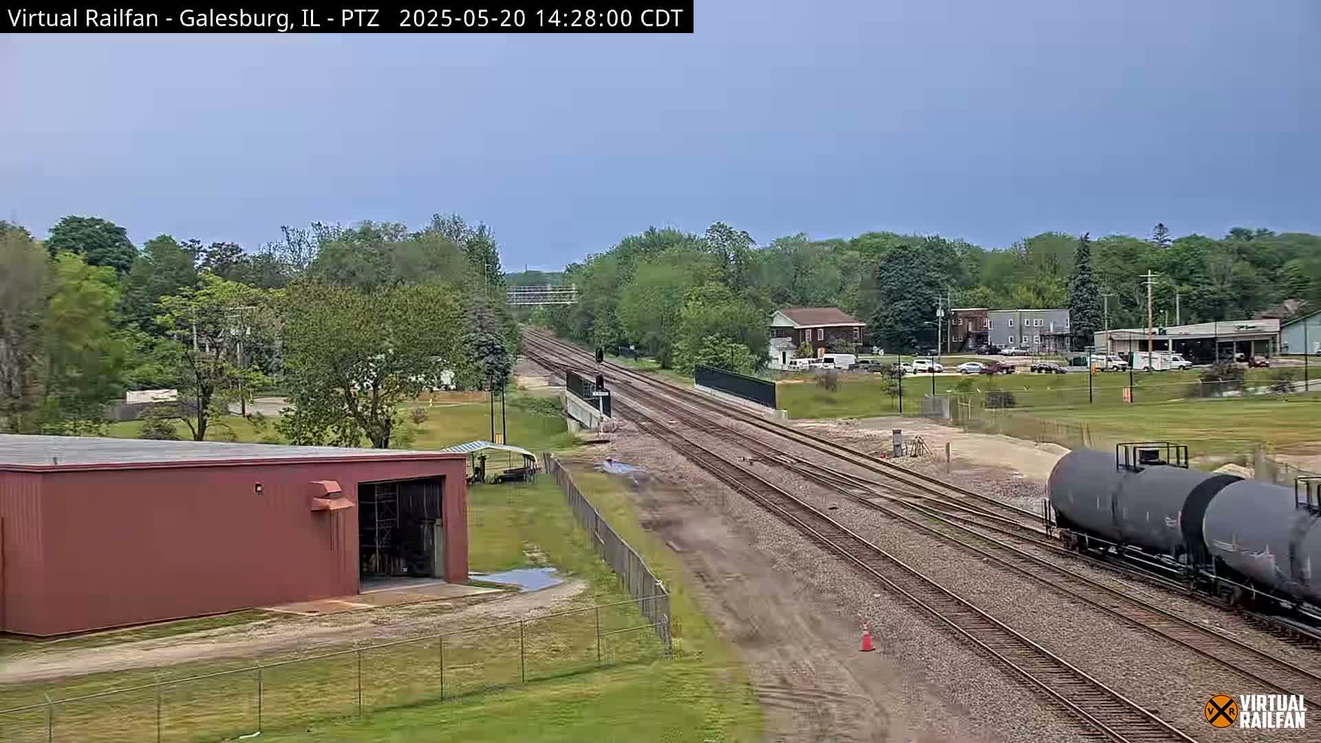 A train of tanker cars moves along railroad tracks that pass through a tree-lined area on an overcast day.