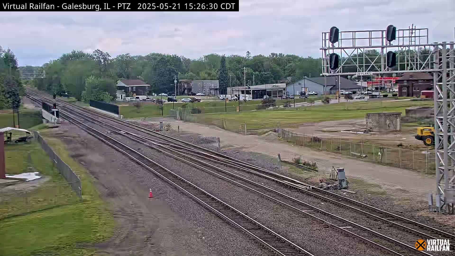 Multiple sets of train tracks extend into the distance under an overcast sky, past a road with buildings and some light construction activity.