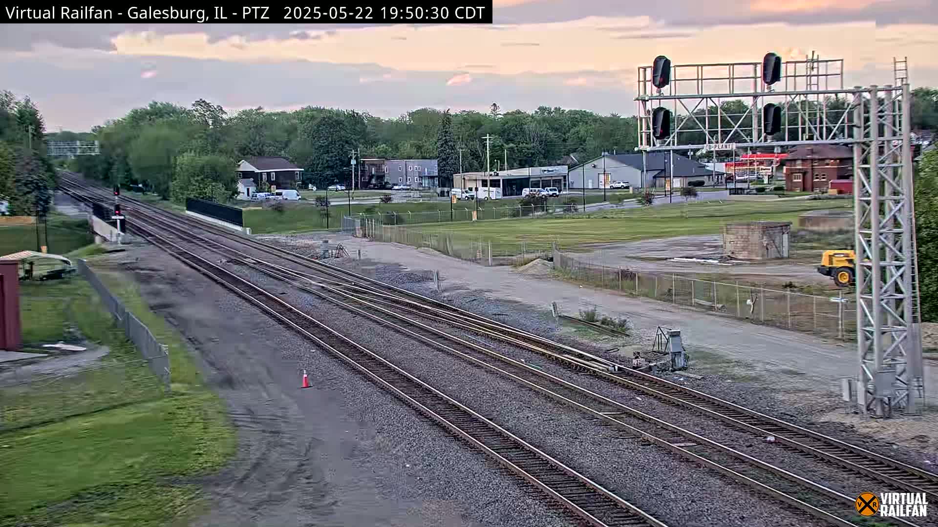 Multiple train tracks extend into the distance under a partly cloudy twilight sky, passing by a small town with industrial areas and a piece of construction equipment visible nearby.