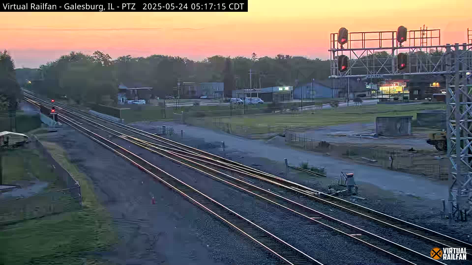 Multiple sets of train tracks stretch into the distance under a sunrise sky, passing near a roadway and buildings.