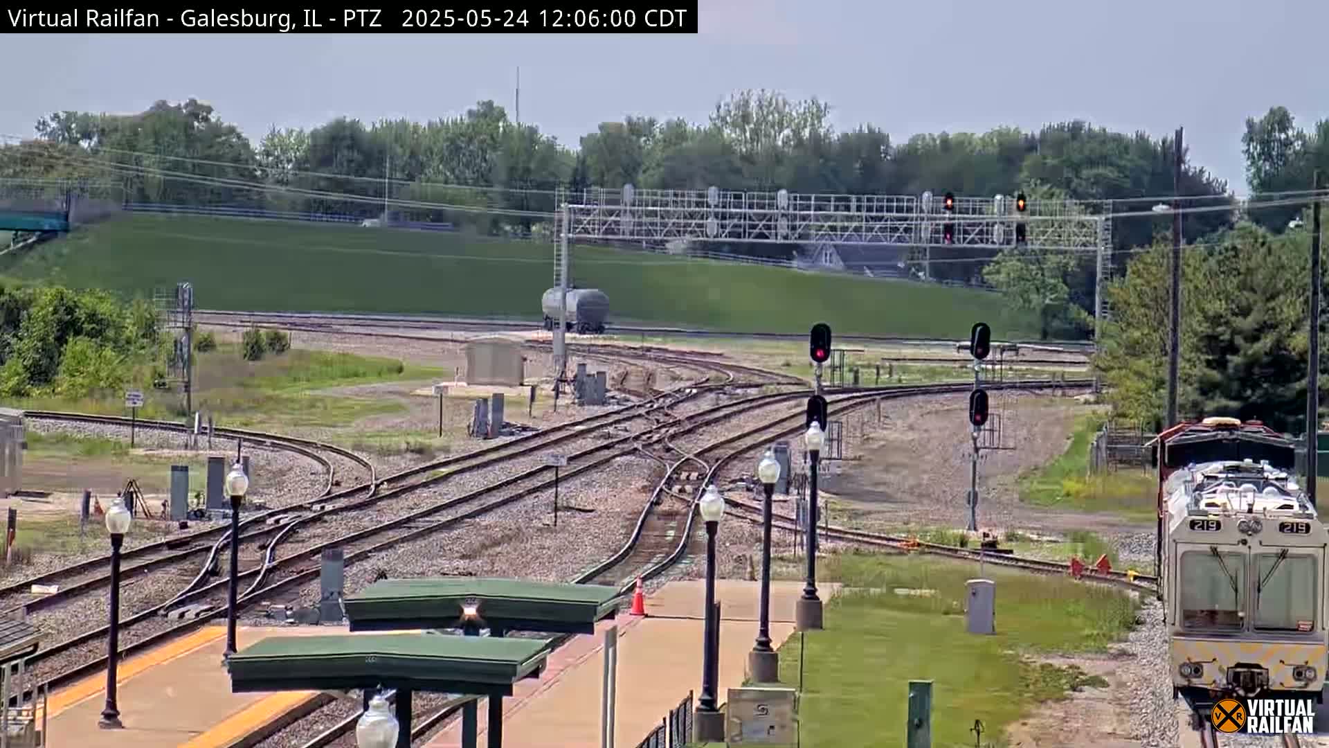 A train approaches a railway junction under a mostly sunny sky, with lush green vegetation surrounding the tracks.
