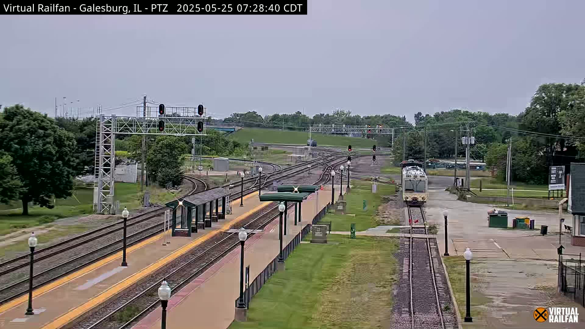 A train approaches a station platform on a partly cloudy day, surrounded by green trees and railway infrastructure.