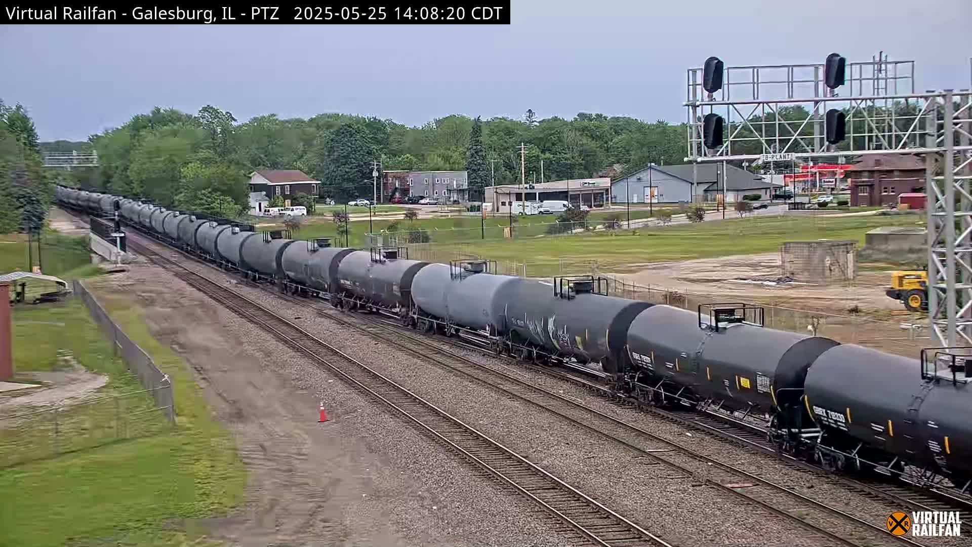 A long train of dark gray tanker cars travels along a railway track on an overcast day, passing by a small town with buildings and some construction equipment visible in the background.