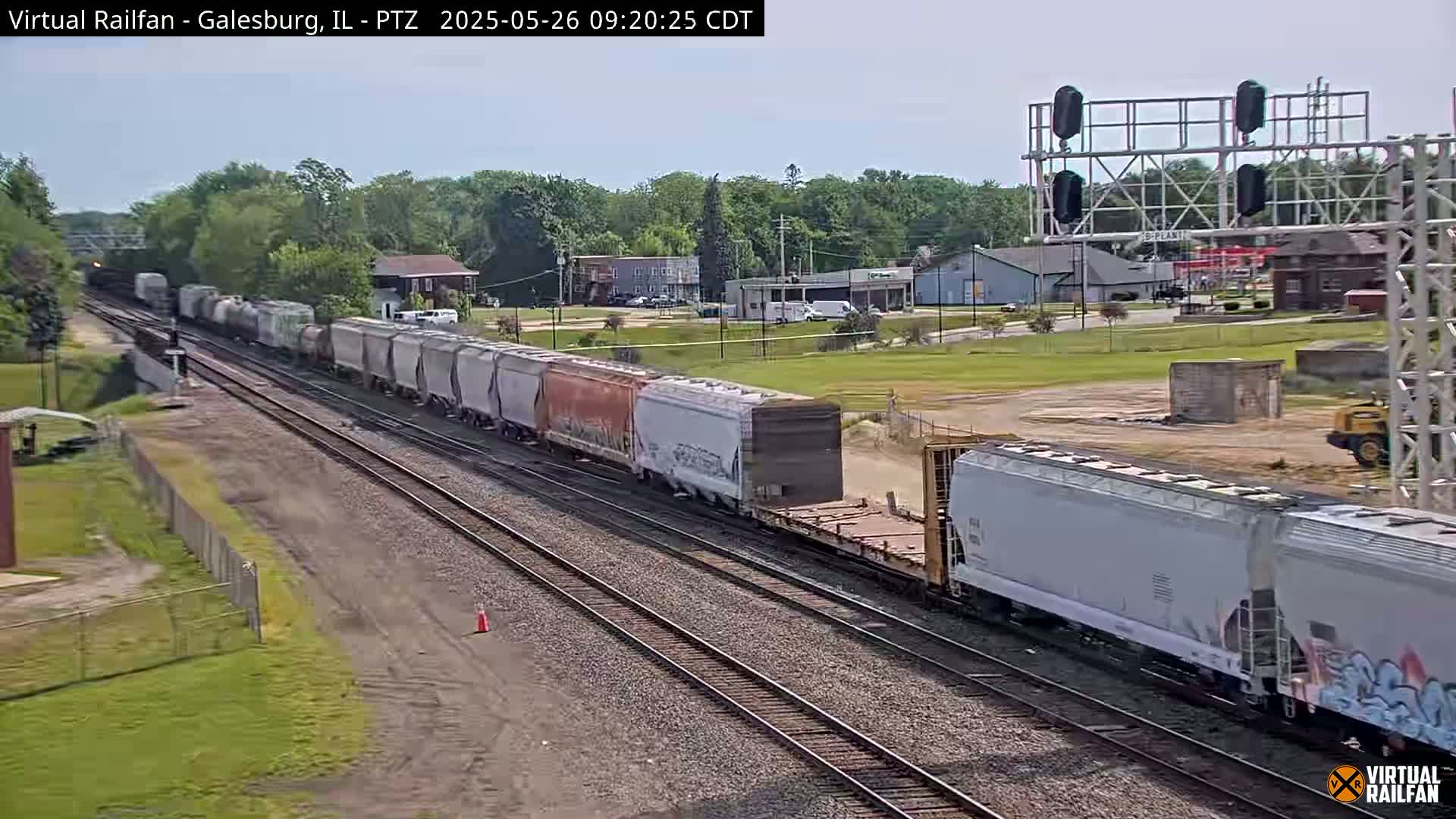 A long freight train passes through a flat, grassy area under a mostly clear sky, near buildings and a railway crossing.