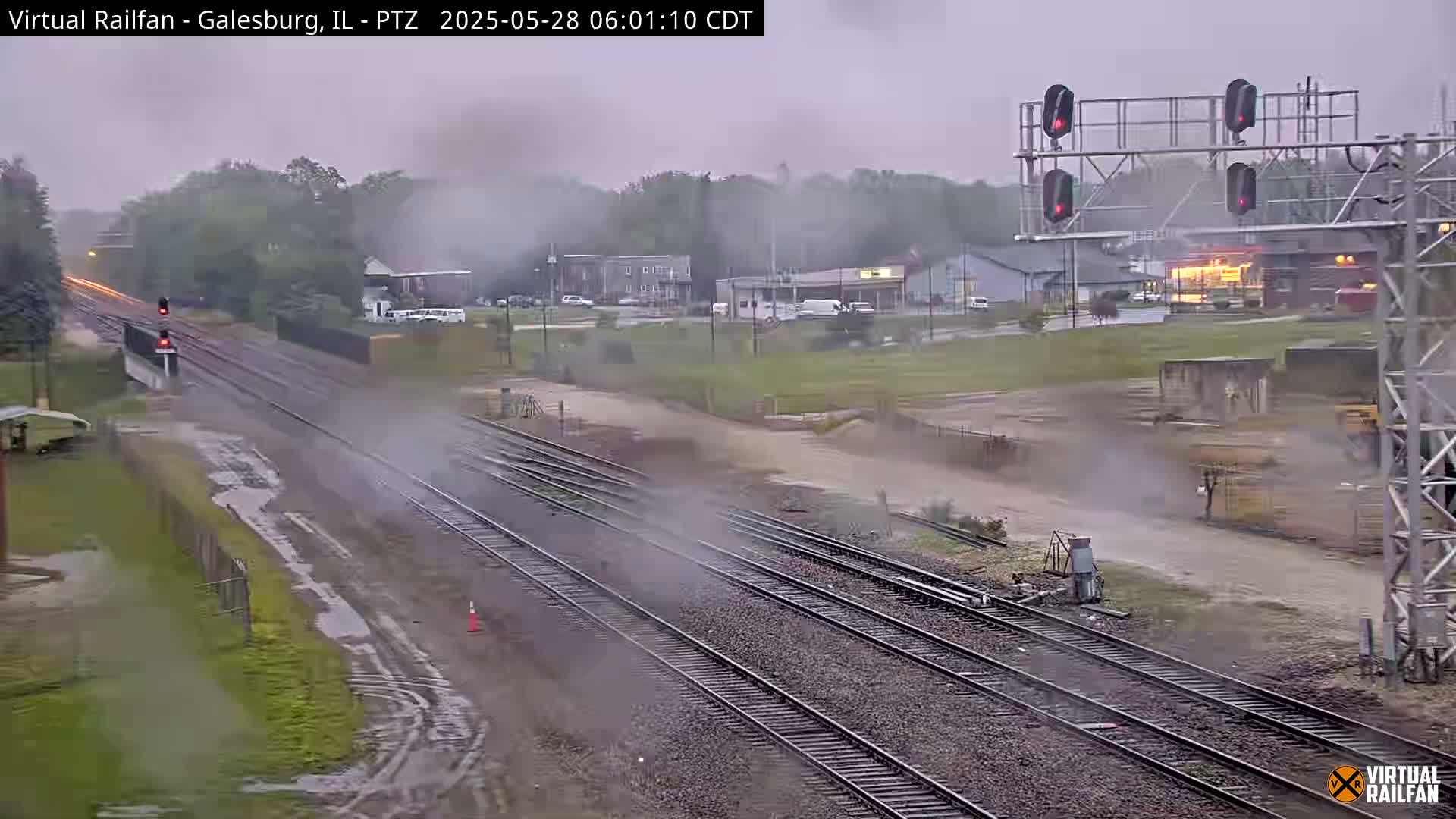 A rain-swept view of multiple railway tracks extending into the distance past a road crossing and a small town under an overcast sky.