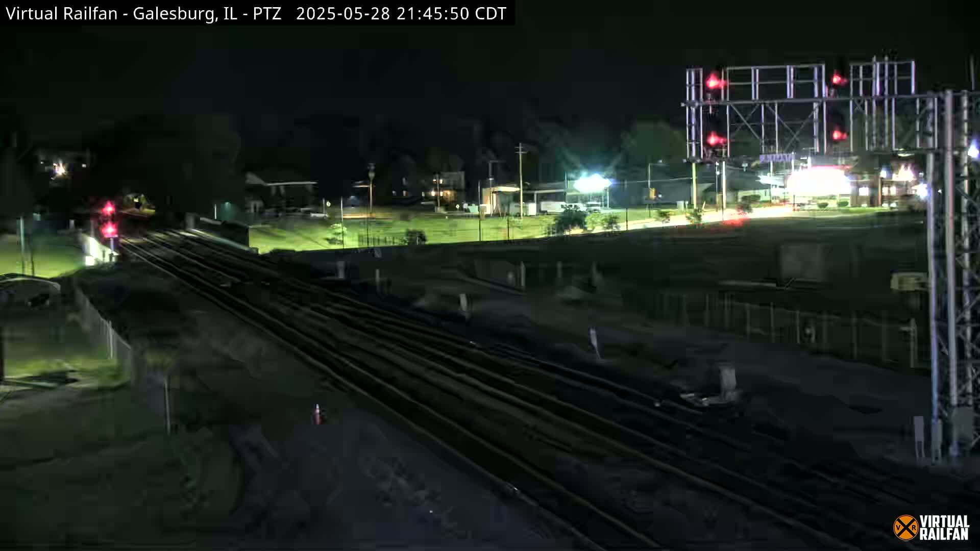 A nighttime view of a railroad crossing with a train approaching,  surrounded by dark, flat land and buildings under a clear sky.