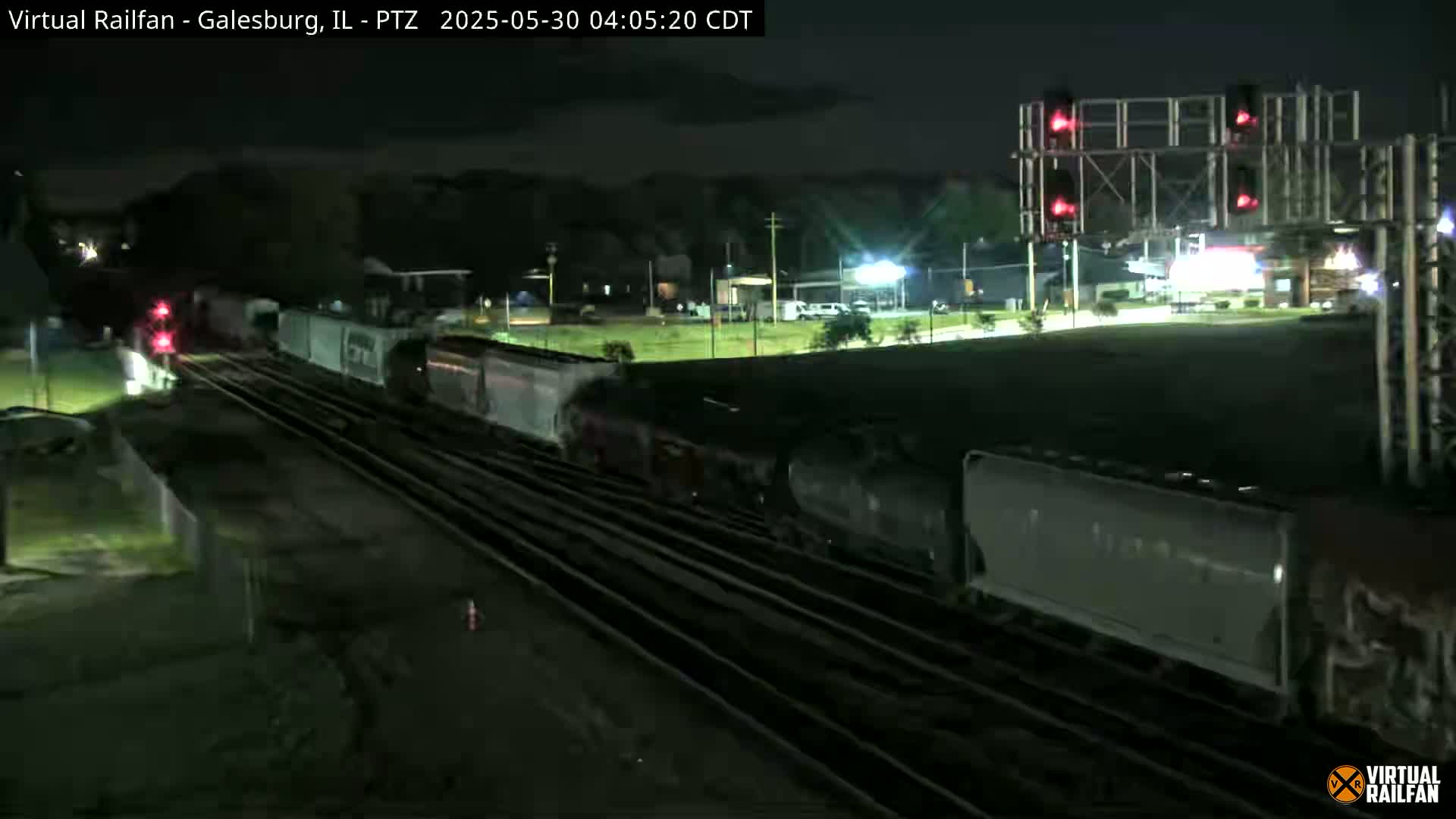 A long freight train passes under red railroad crossing lights at night.