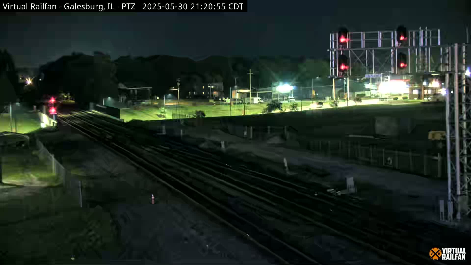 A nighttime view of railroad tracks and a train passing under a railway crossing signal, with a grassy area and buildings visible in the background under a clear sky.