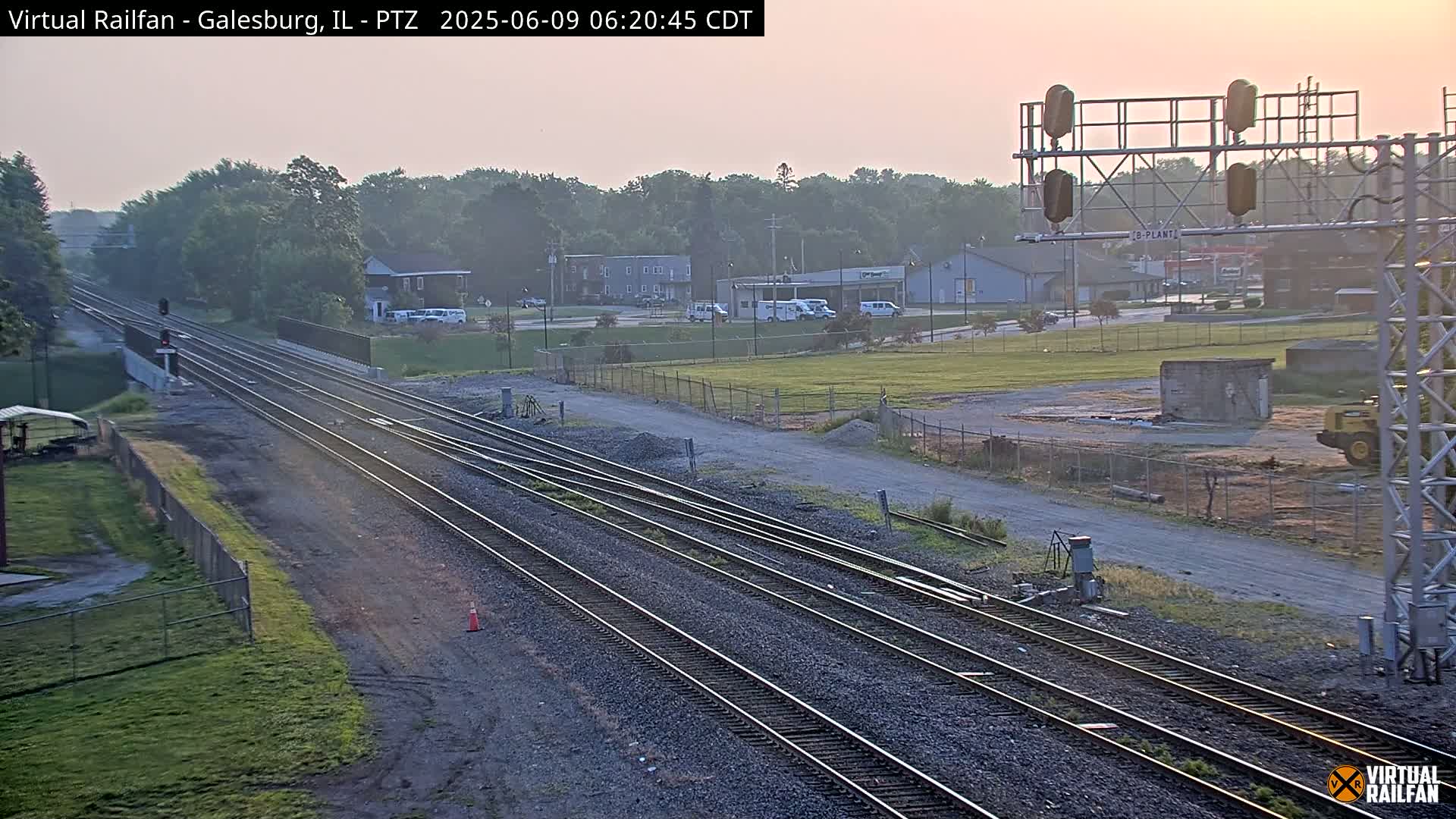 Multiple sets of train tracks stretch into the distance under a hazy, sunrise sky, passing by a small town with buildings and vehicles.