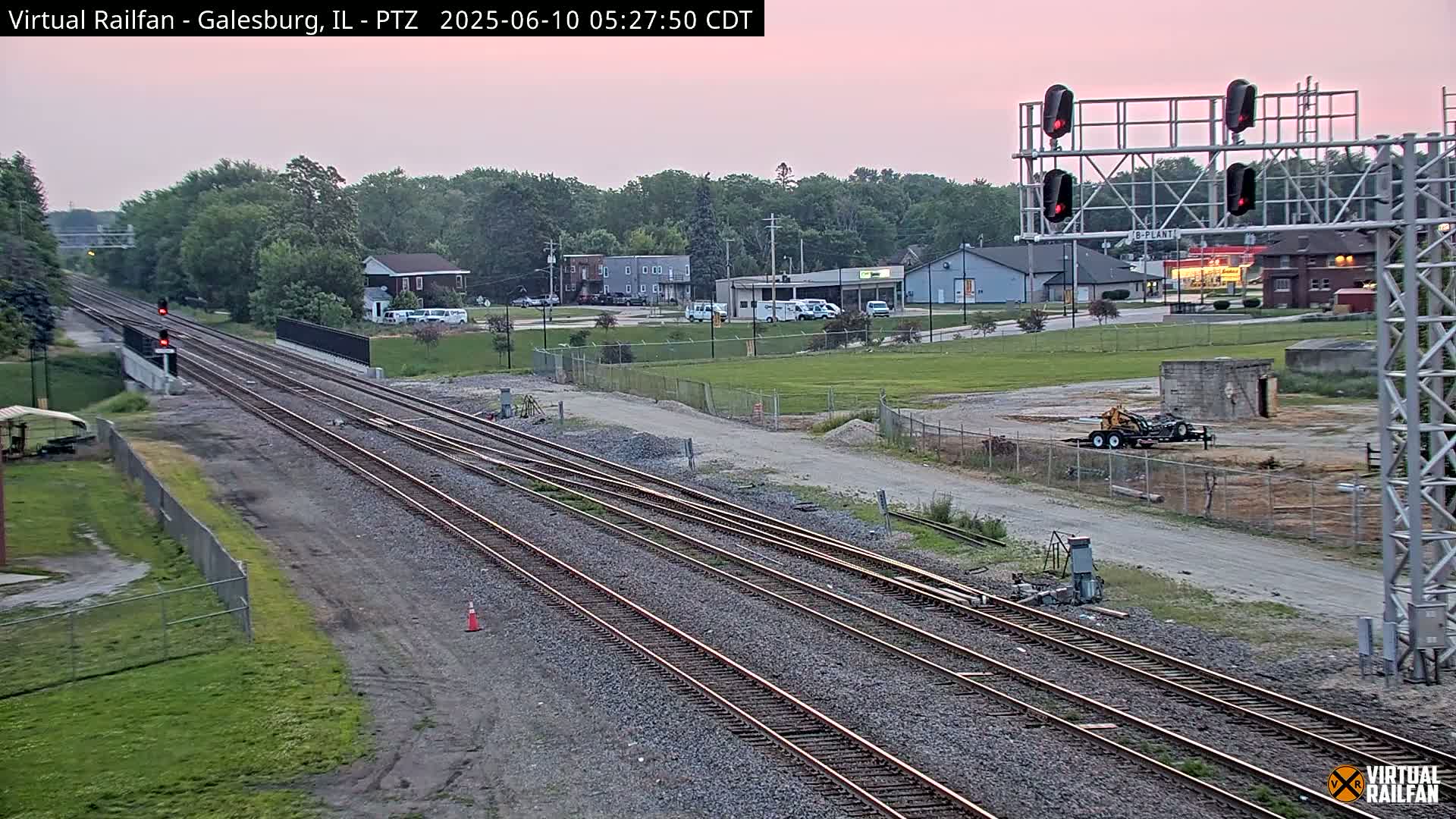 Multiple sets of train tracks extend into the distance under a dusky pink sky, passing by buildings and a grassy area.