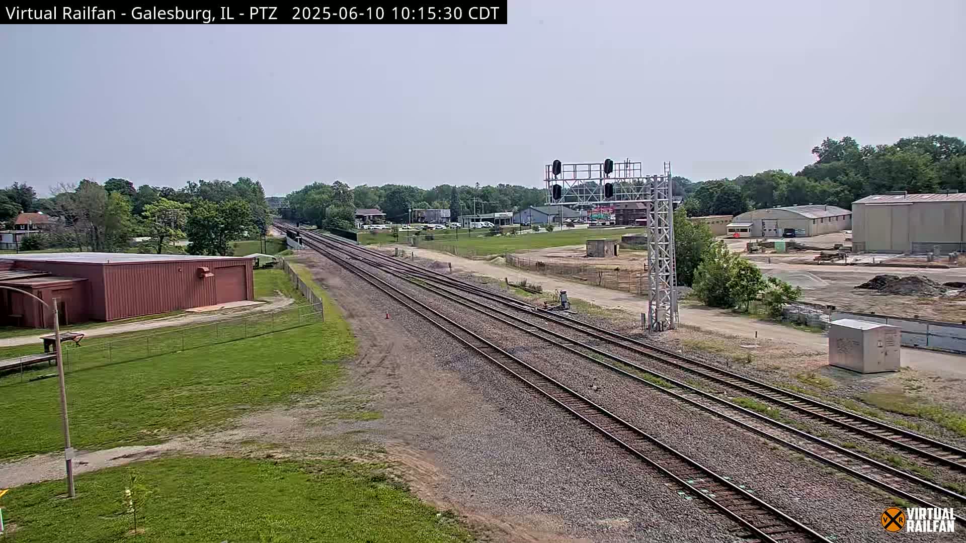 Multiple sets of train tracks extend into the distance under a mostly clear sky, passing by industrial buildings and grassy areas.
