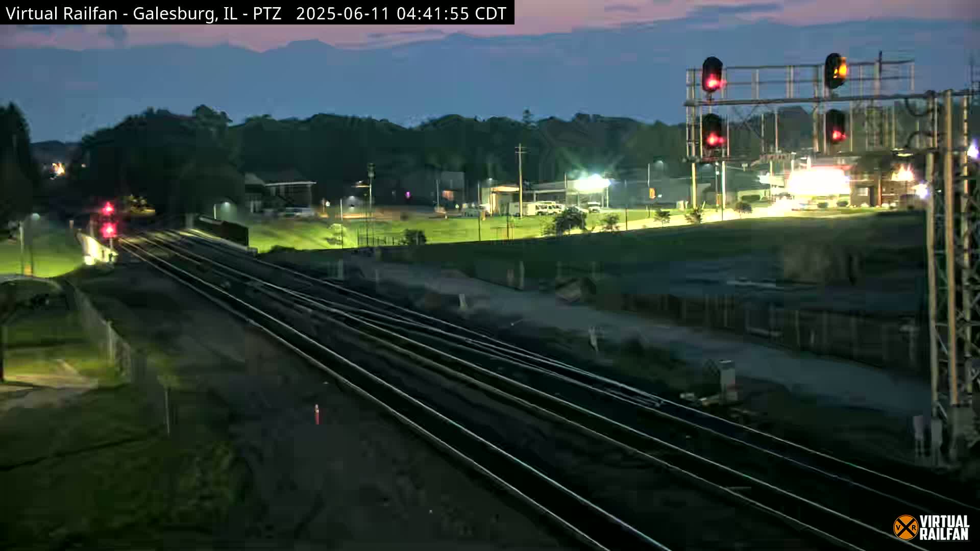 A nighttime view of several sets of train tracks extending into the distance, passing under a railway crossing signal, with a dimly lit landscape and trees in the background under a twilight sky.