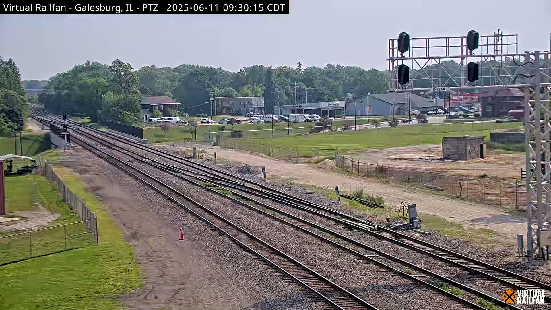 Multiple sets of train tracks extend into the distance under a partly sunny sky, passing by a road with buildings and cars visible in the background.