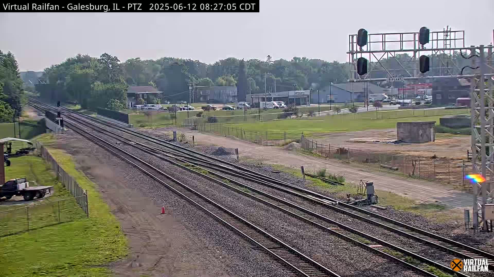 Multiple sets of train tracks extend into the distance under a clear sky, passing in front of a small town with some light industrial buildings.
