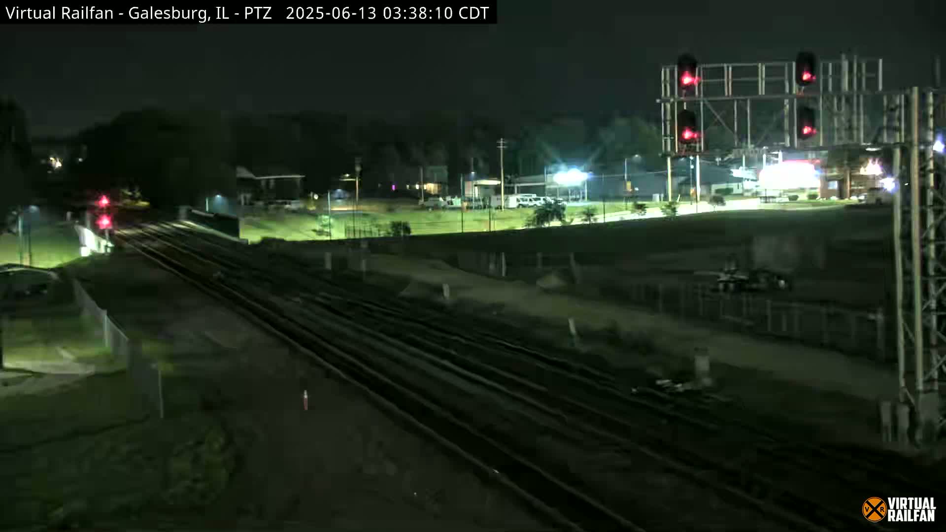 A nighttime view of railroad tracks and a crossing with red signal lights, showing a train approaching in the distance.