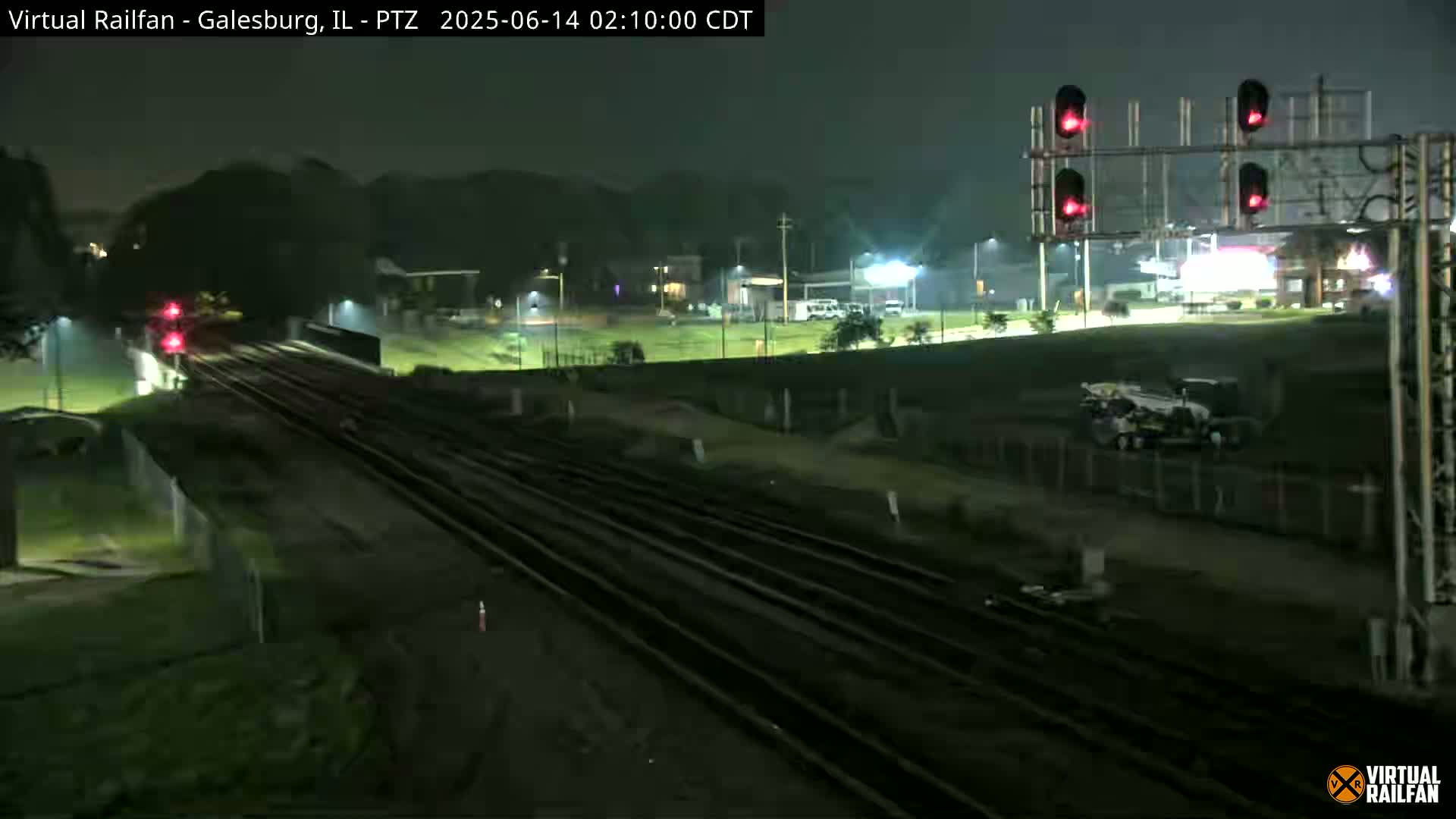 A nighttime view of railroad tracks intersecting with a road, featuring red traffic signals, some surrounding buildings, and construction equipment.