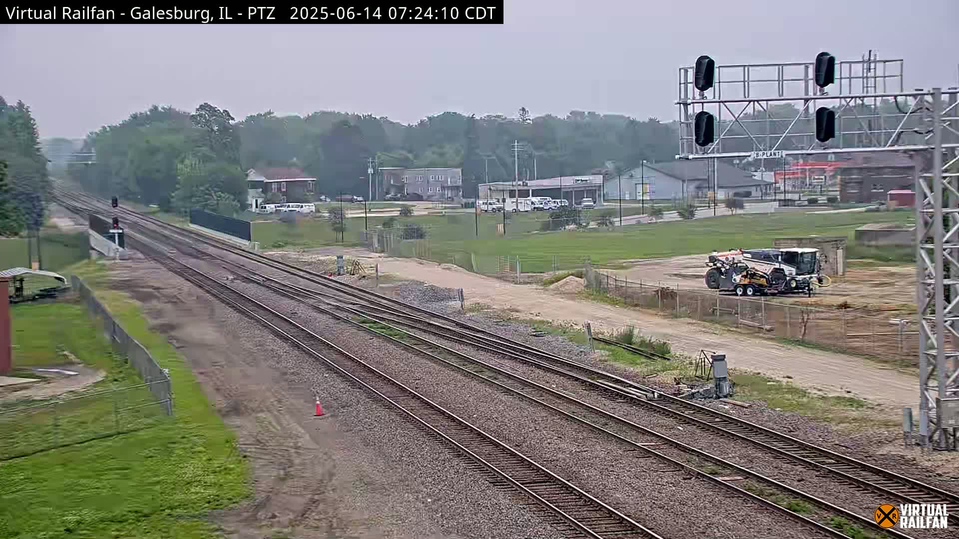 Multiple sets of train tracks stretch into the distance under an overcast sky, with a piece of construction equipment visible in the middle ground and a few buildings in the background.