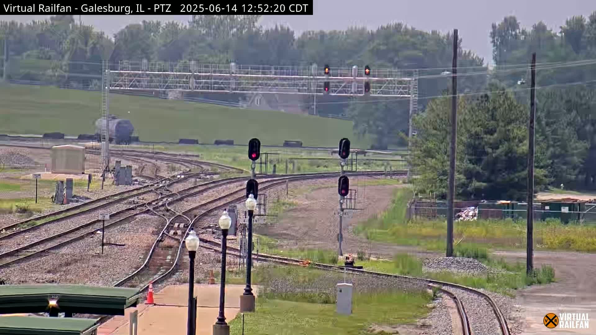 A sunny outdoor scene shows a railroad yard with multiple train tracks curving and intersecting, several signal lights, and some vegetation.