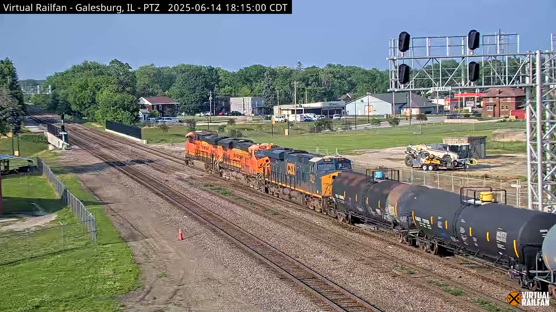 A sunny outdoor scene shows a railroad yard with multiple train tracks curving and intersecting, several signal lights, and some vegetation.