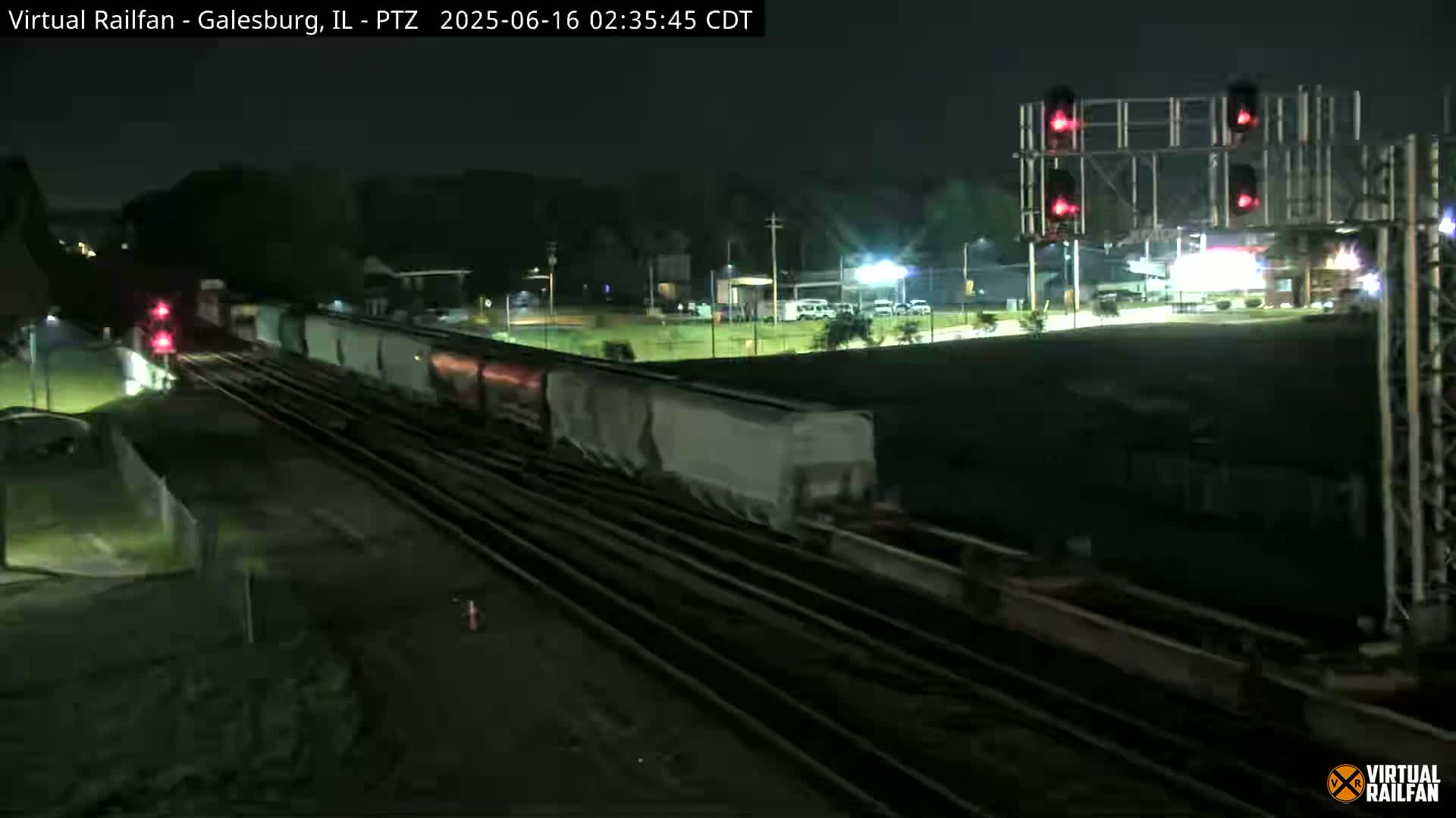 At night, a long freight train passes under a railway crossing signal with red lights illuminated.