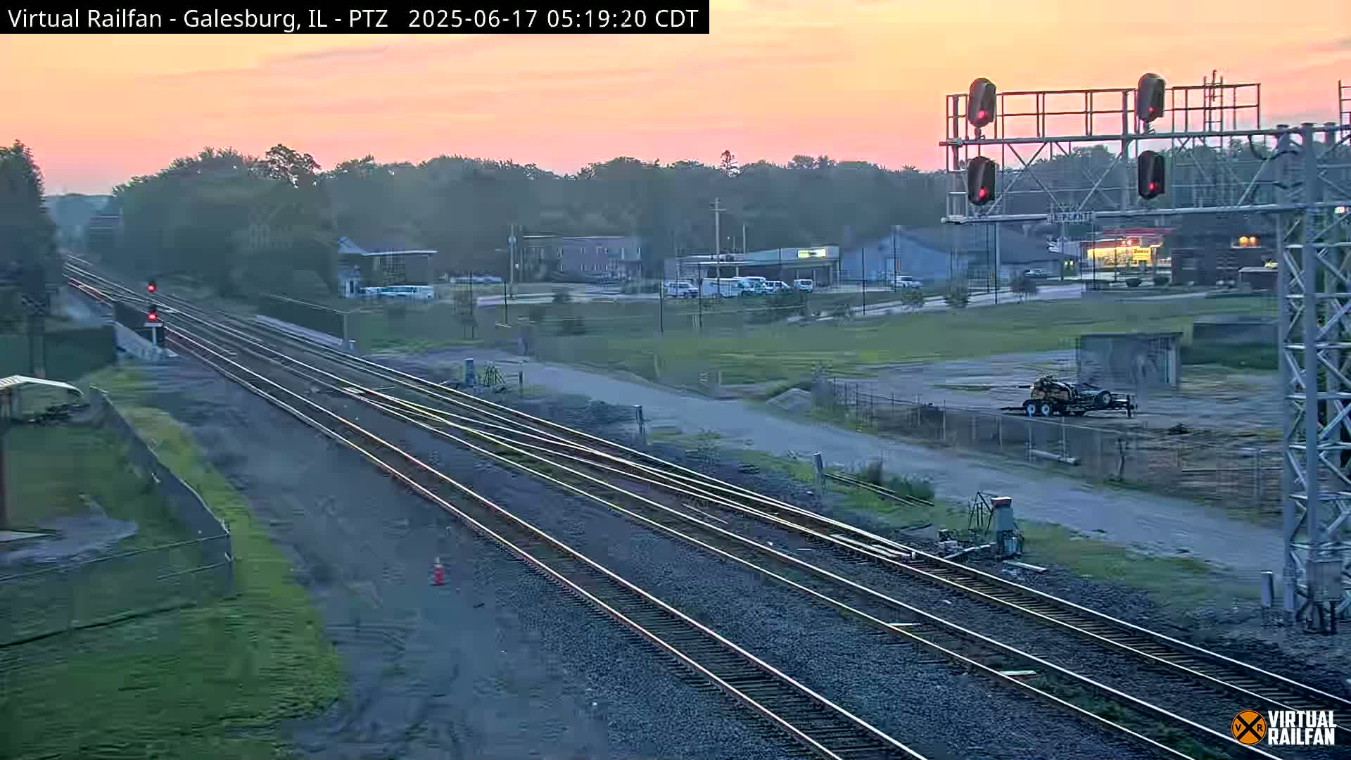 Multiple train tracks extend into the distance under a pinkish-orange sunrise sky, with a few buildings and vehicles visible in the background.
