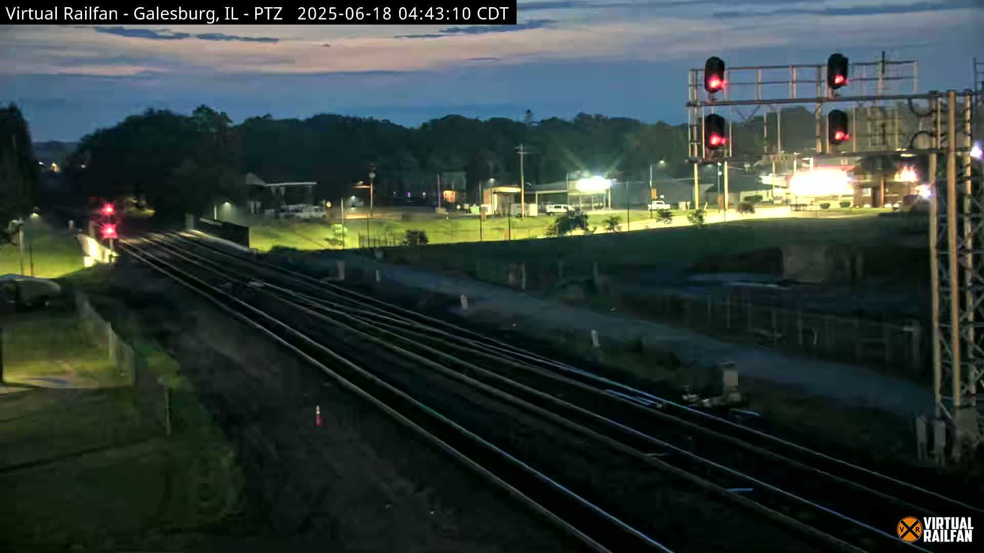 A nighttime view of multiple train tracks extending into the distance, under a twilight sky, with a road and buildings visible in the background.