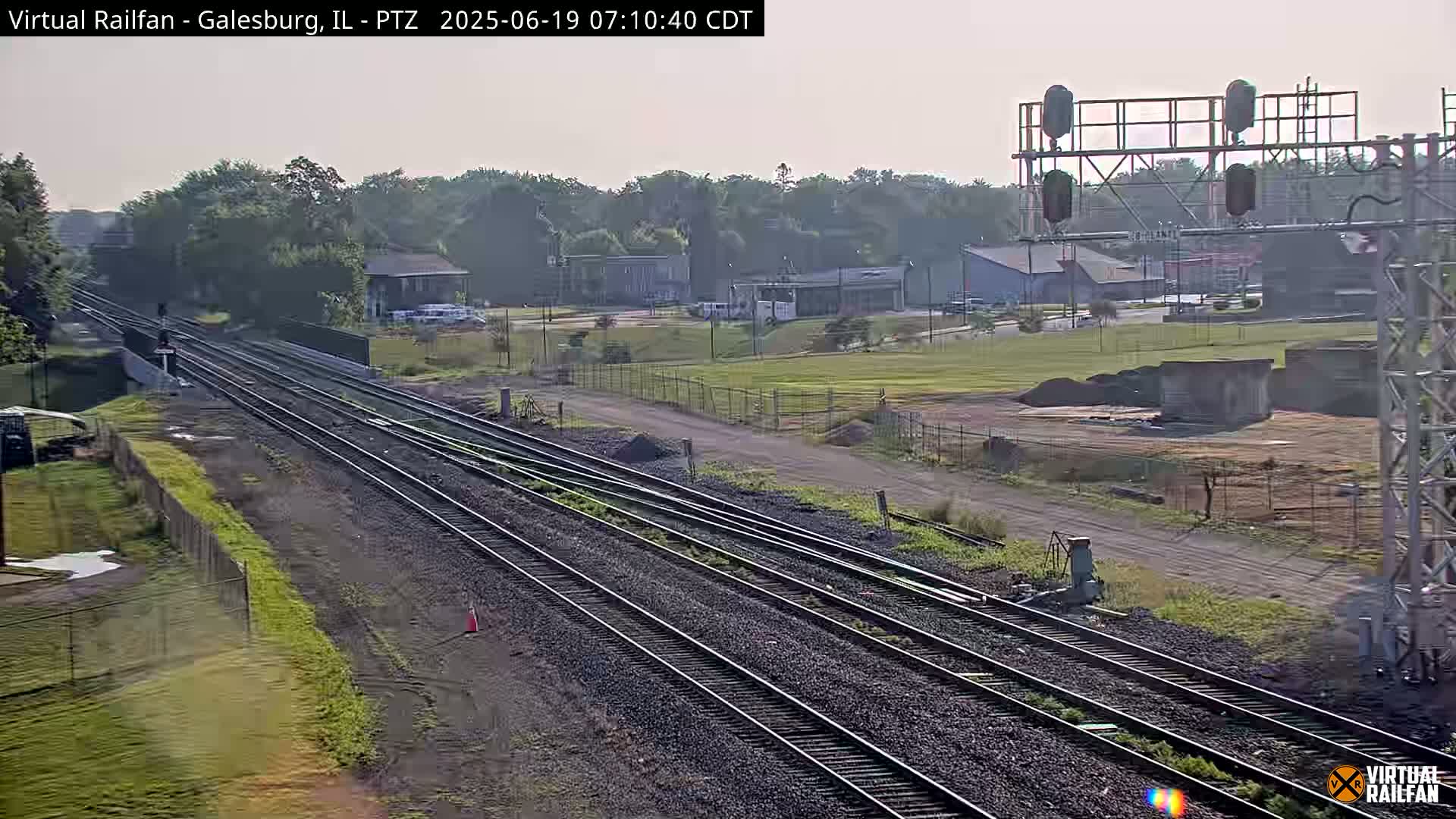 Multiple sets of train tracks stretch into the distance on a mostly clear day, passing in front of a low-lying industrial area and some trees.