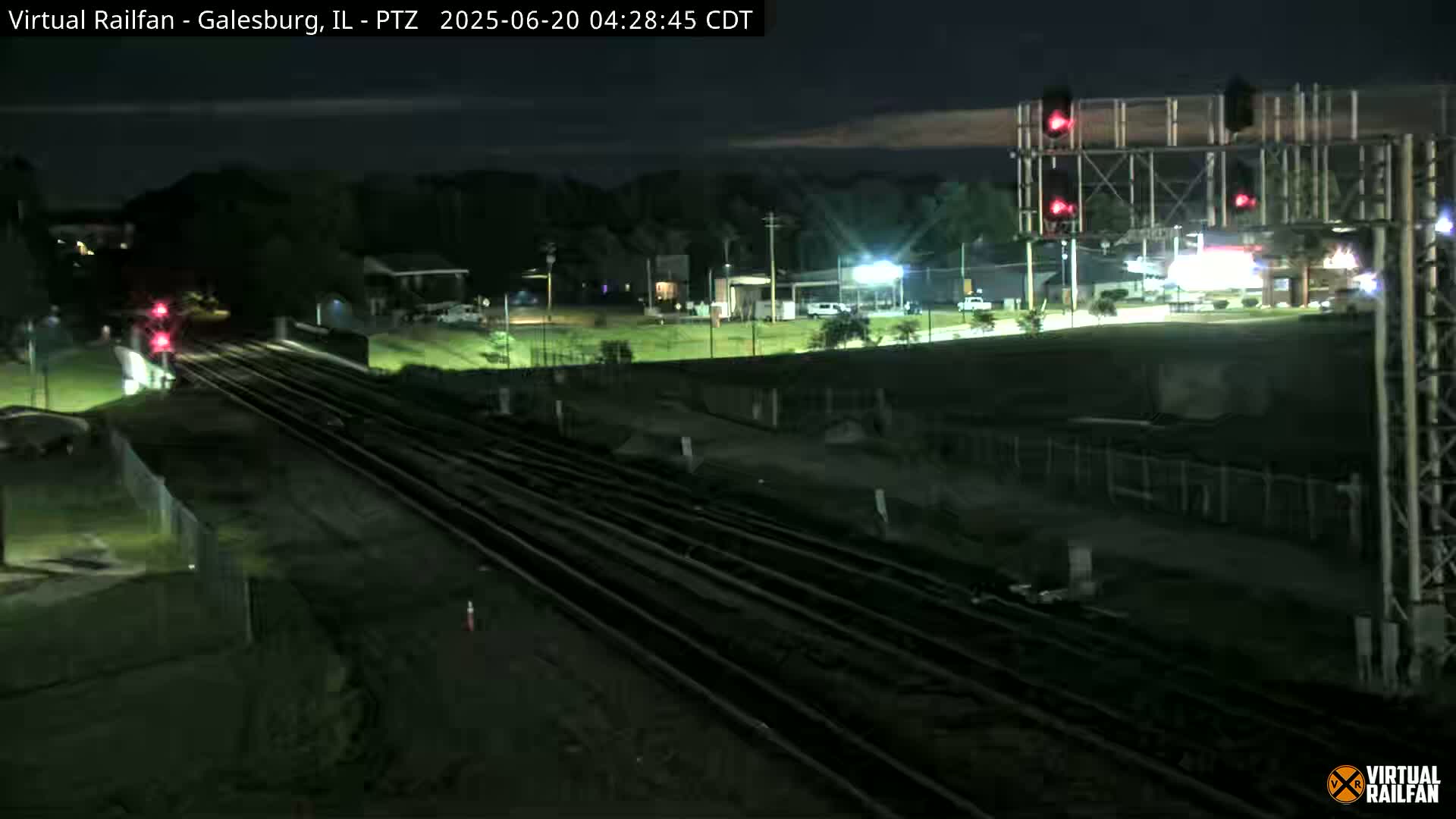 At night, multiple train tracks extend into the distance, passing under a road and near a fenced area with red traffic signals visible.