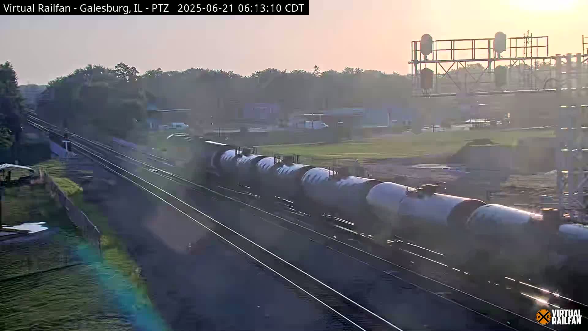 A long freight train of tanker cars is traveling on railroad tracks at sunrise in hazy conditions.