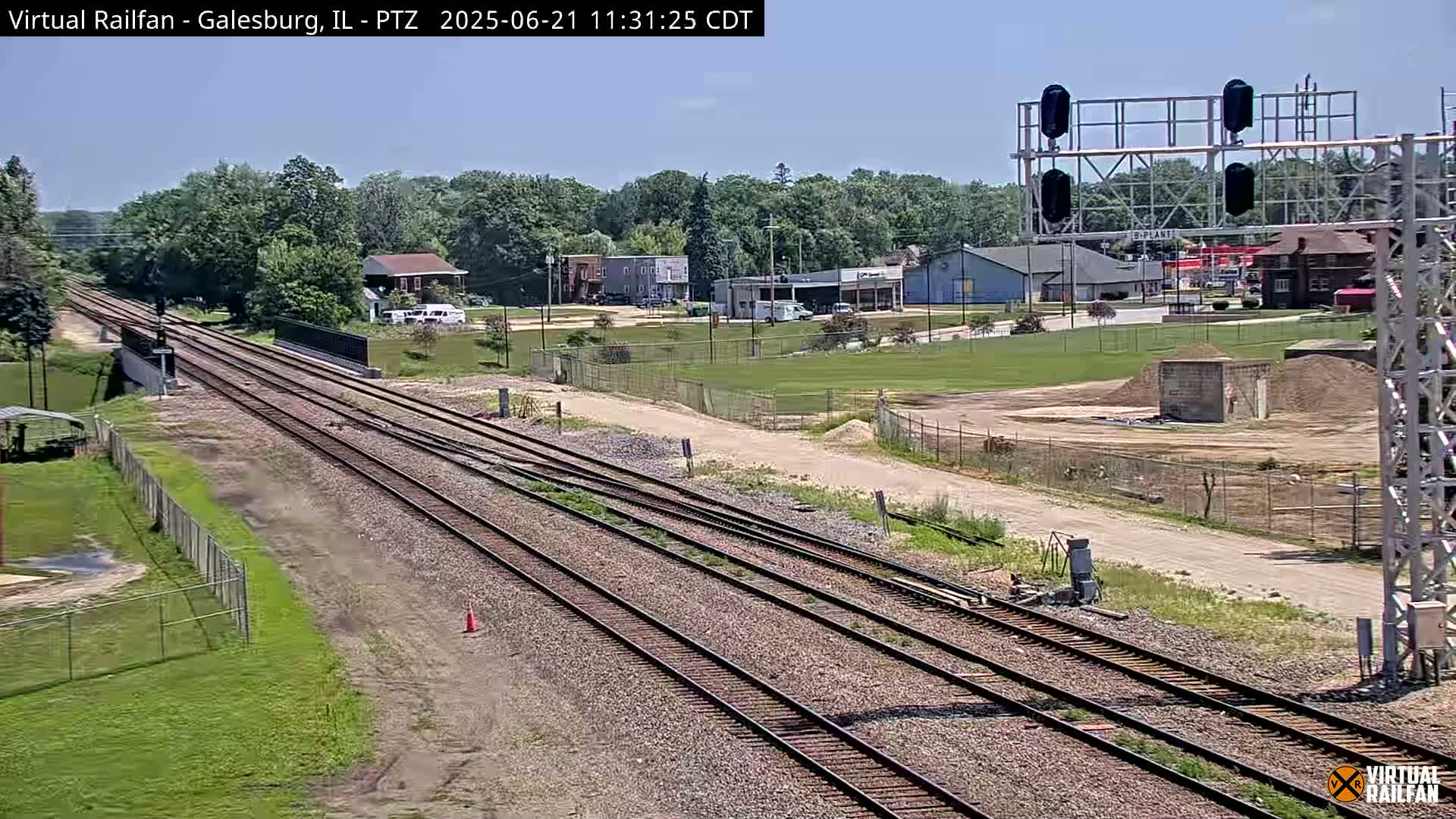 Multiple train tracks stretch toward a small town under a clear sky, with some construction visible near a grassy field.
