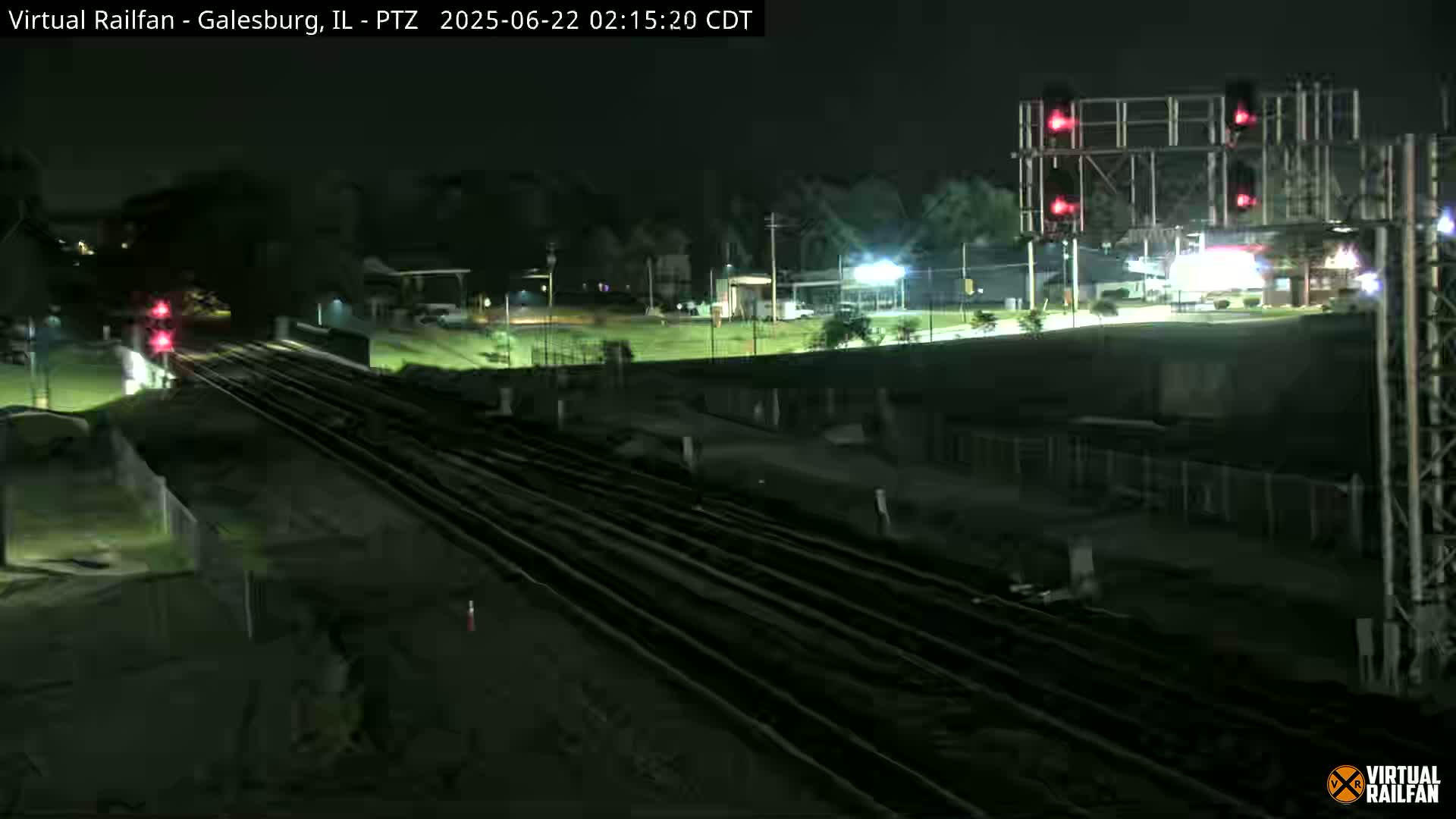 A nighttime view of railroad tracks and a road crossing under illuminated traffic signals, with some surrounding buildings and land visible in the dark.