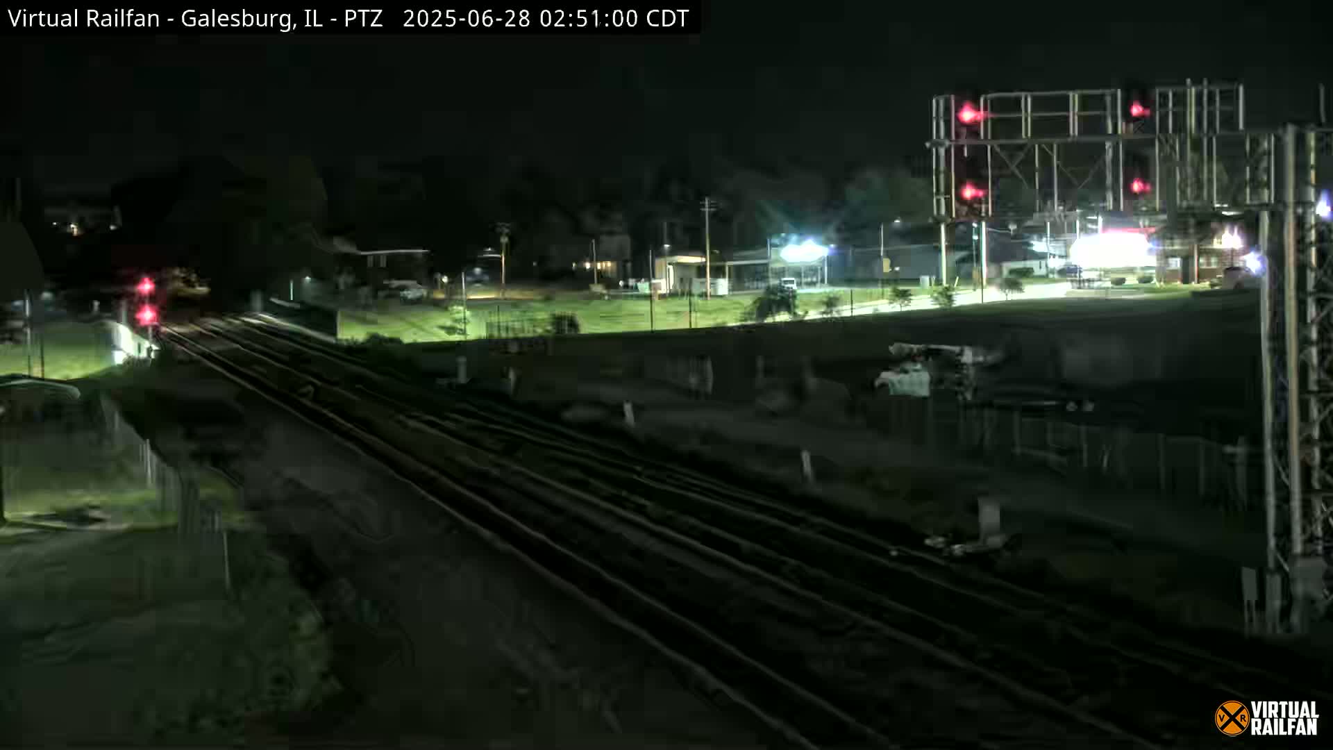 A nighttime view of railroad tracks, crossing signals, and a surrounding area with sparse vegetation and buildings under a clear sky.