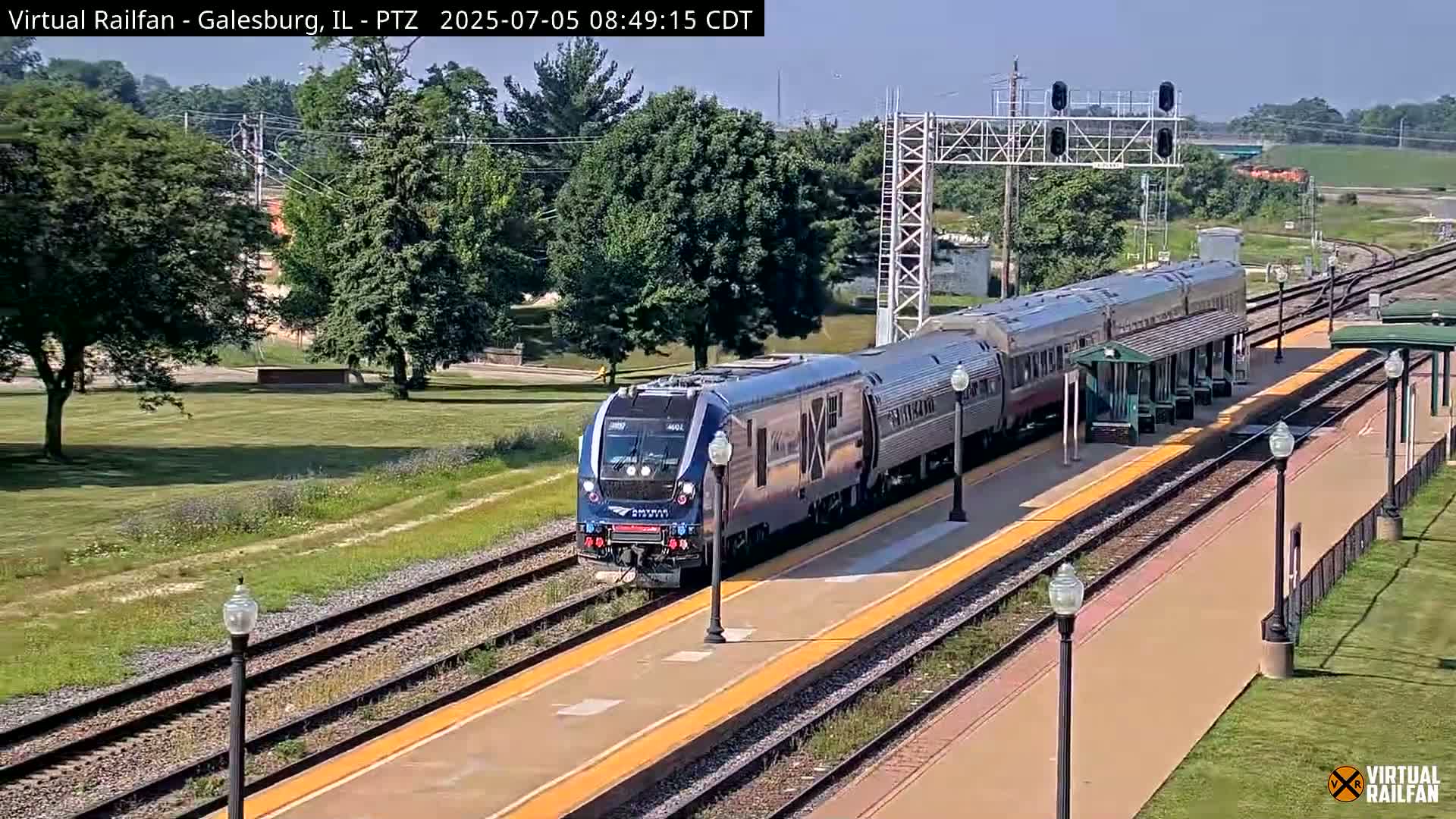 A long, blue and silver passenger train is arriving at a station on a sunny day.