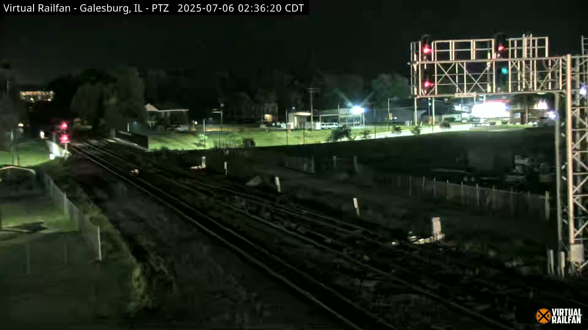 A nighttime view of railroad tracks and surrounding landscape, with a train partially visible in the distance under dark, clear skies.