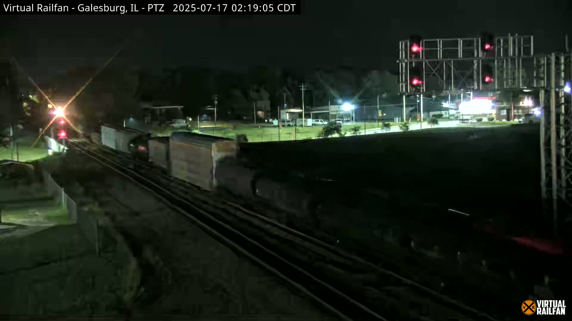 At night, a long freight train passes under a set of red railroad crossing lights.