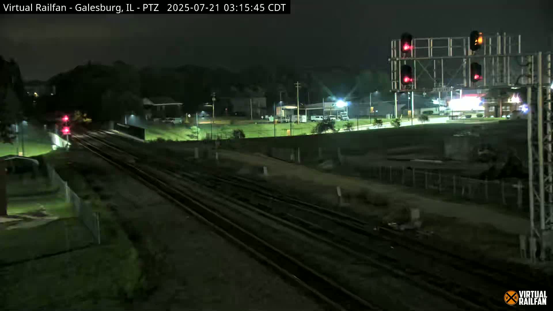 A nighttime view of railroad tracks and a nearby road, with traffic signals visible, under clear skies.