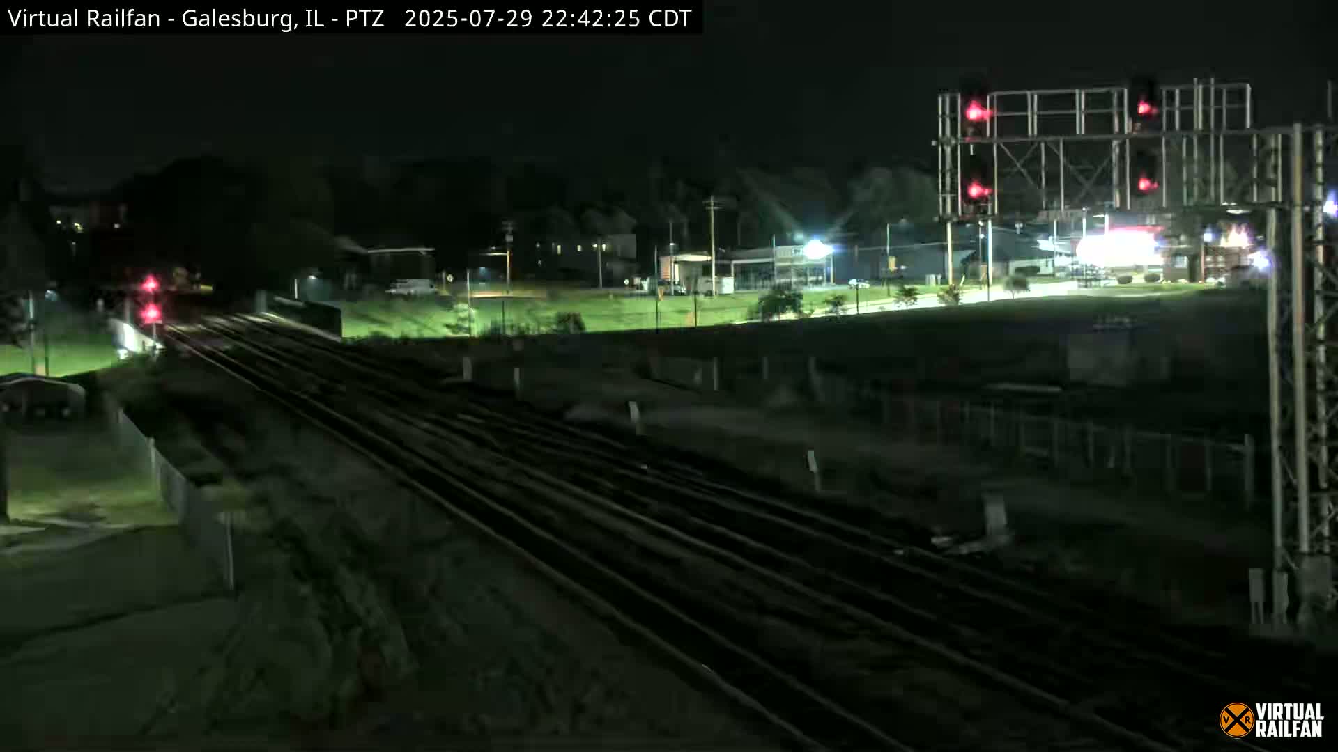 A nighttime view of railroad tracks and a nearby road, with traffic signals visible, under clear skies.