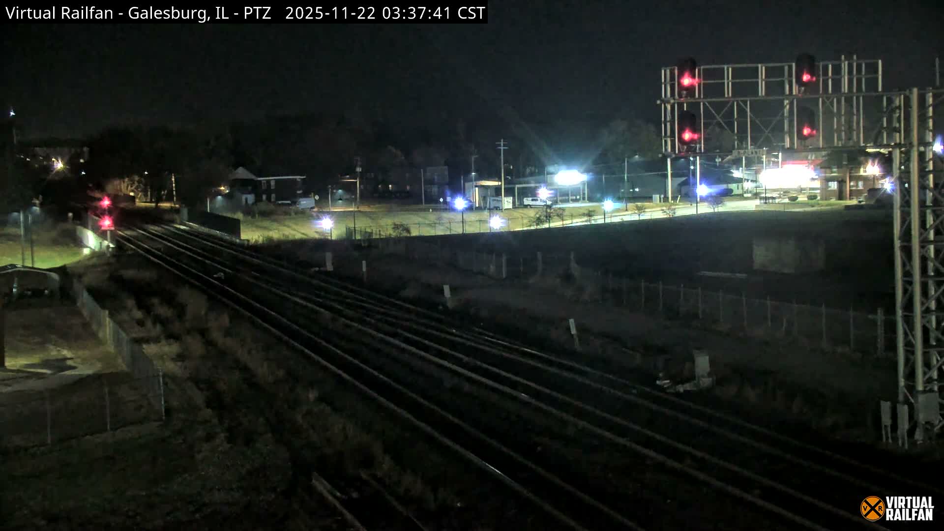 Multiple parallel train tracks with active red signal lights stretch into the distance at night under a clear sky, illuminated by distant artificial lighting from surrounding buildings and streetlights.