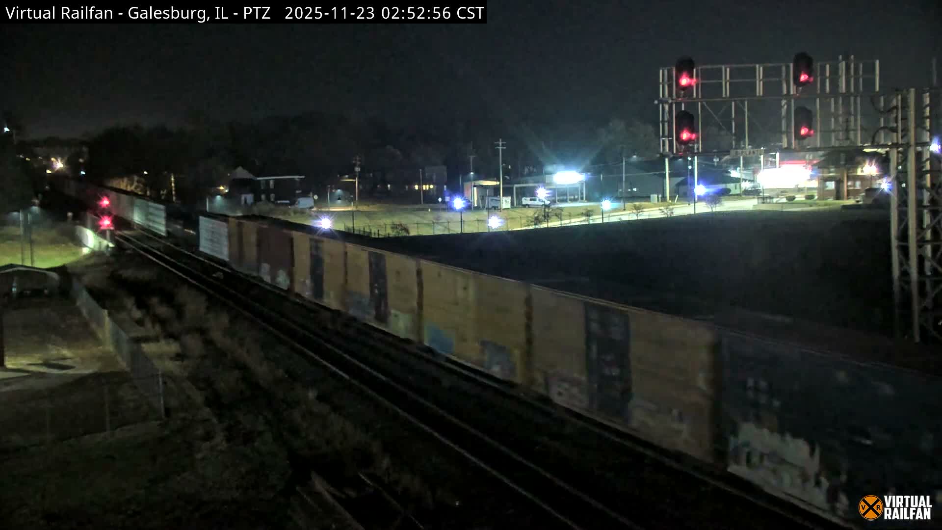 A long freight train with diverse cars travels along multiple tracks at night under clear skies, with red signal lights illuminated on an overhead gantry and various distant lights from buildings and streetlights.
