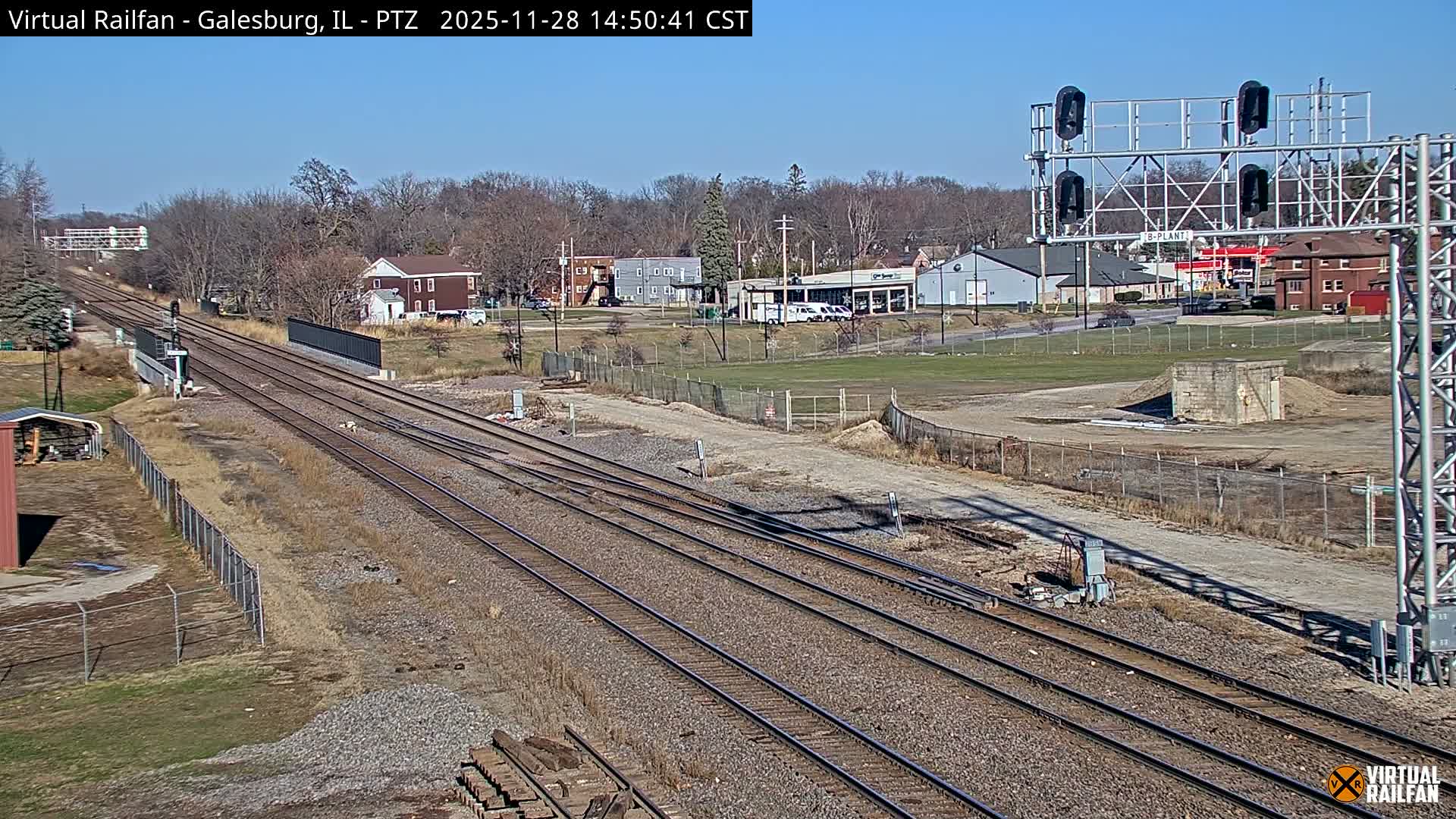 Multiple railway tracks converge and extend through an area with bare trees and various buildings under a clear, sunny sky.
