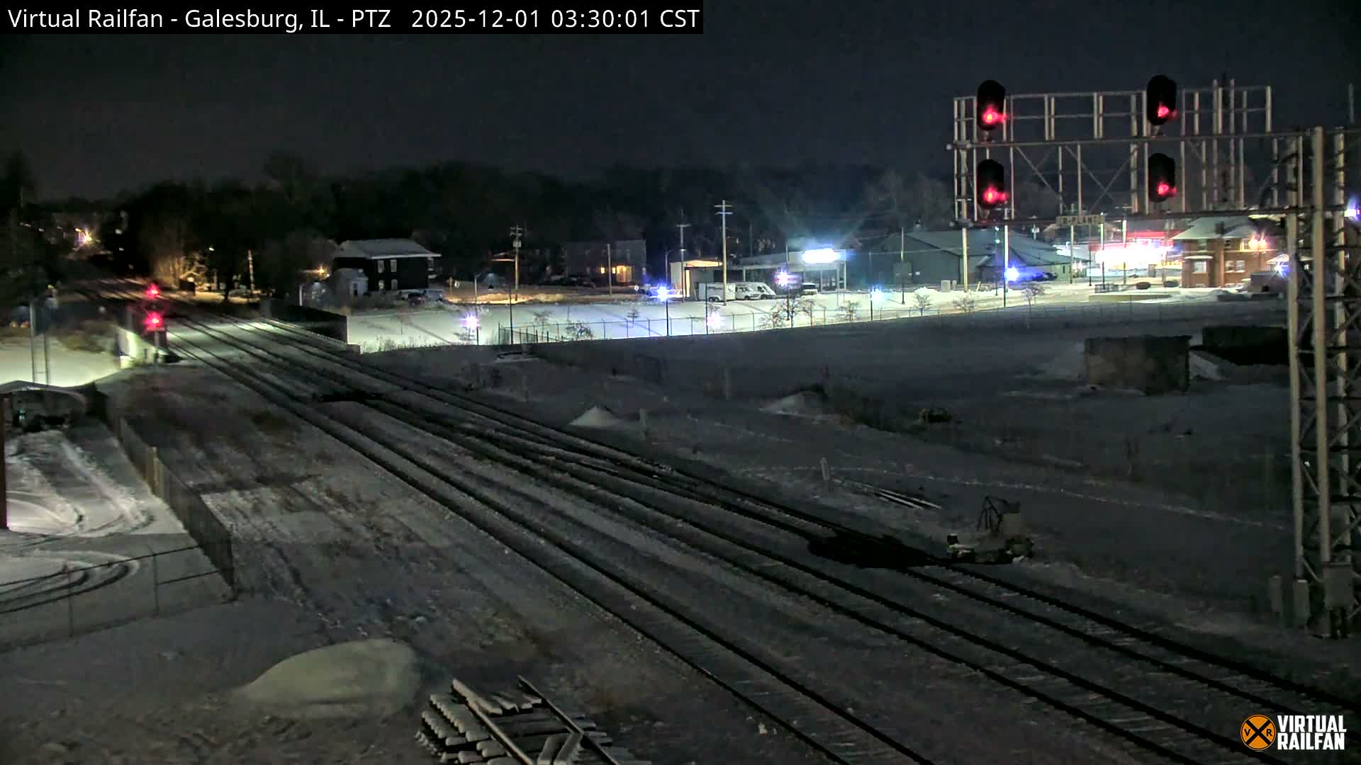 A wintry night scene depicts numerous snow-dusted train tracks stretching into the distance, illuminated by multiple bright red railway signals and distant urban lights under a clear sky.