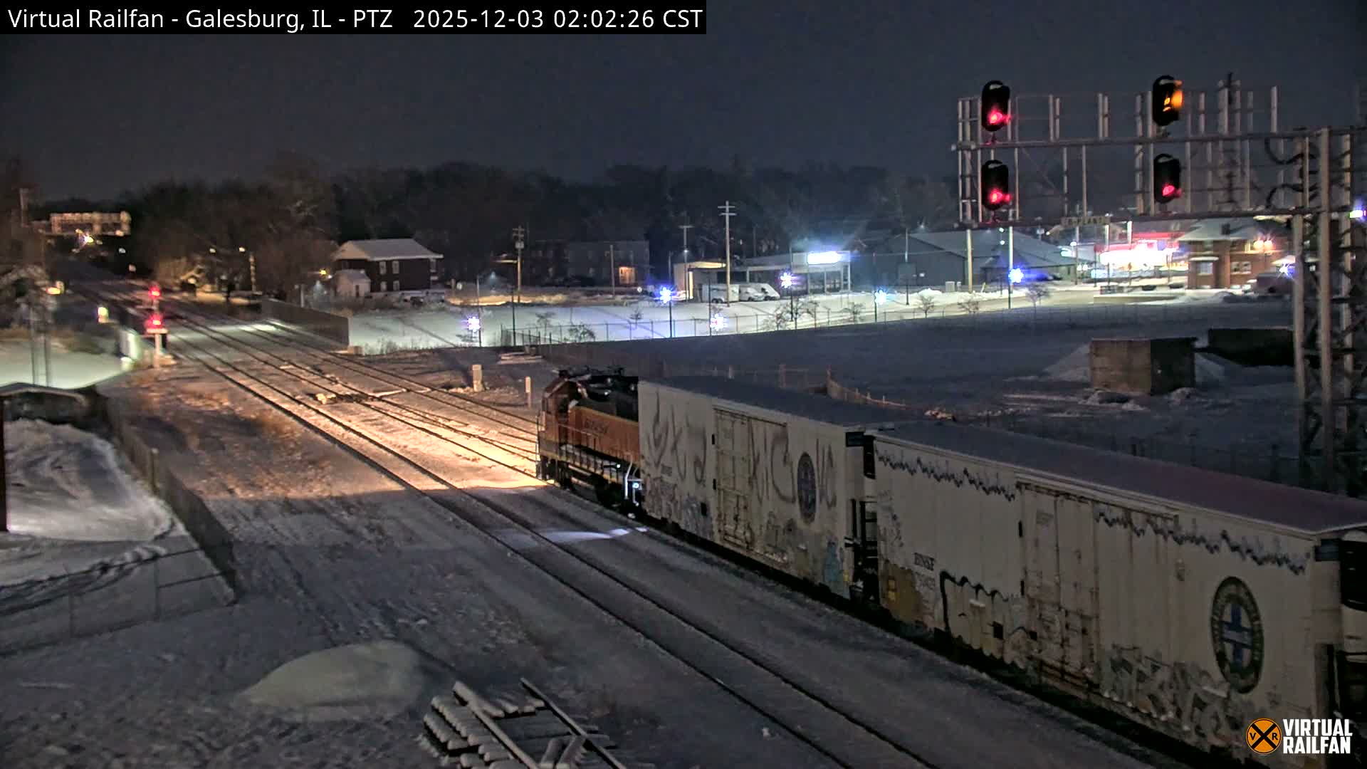 A freight train travels on snow-covered tracks at night, illuminated by its headlight and various distant city lights and railway signals in an urban setting.