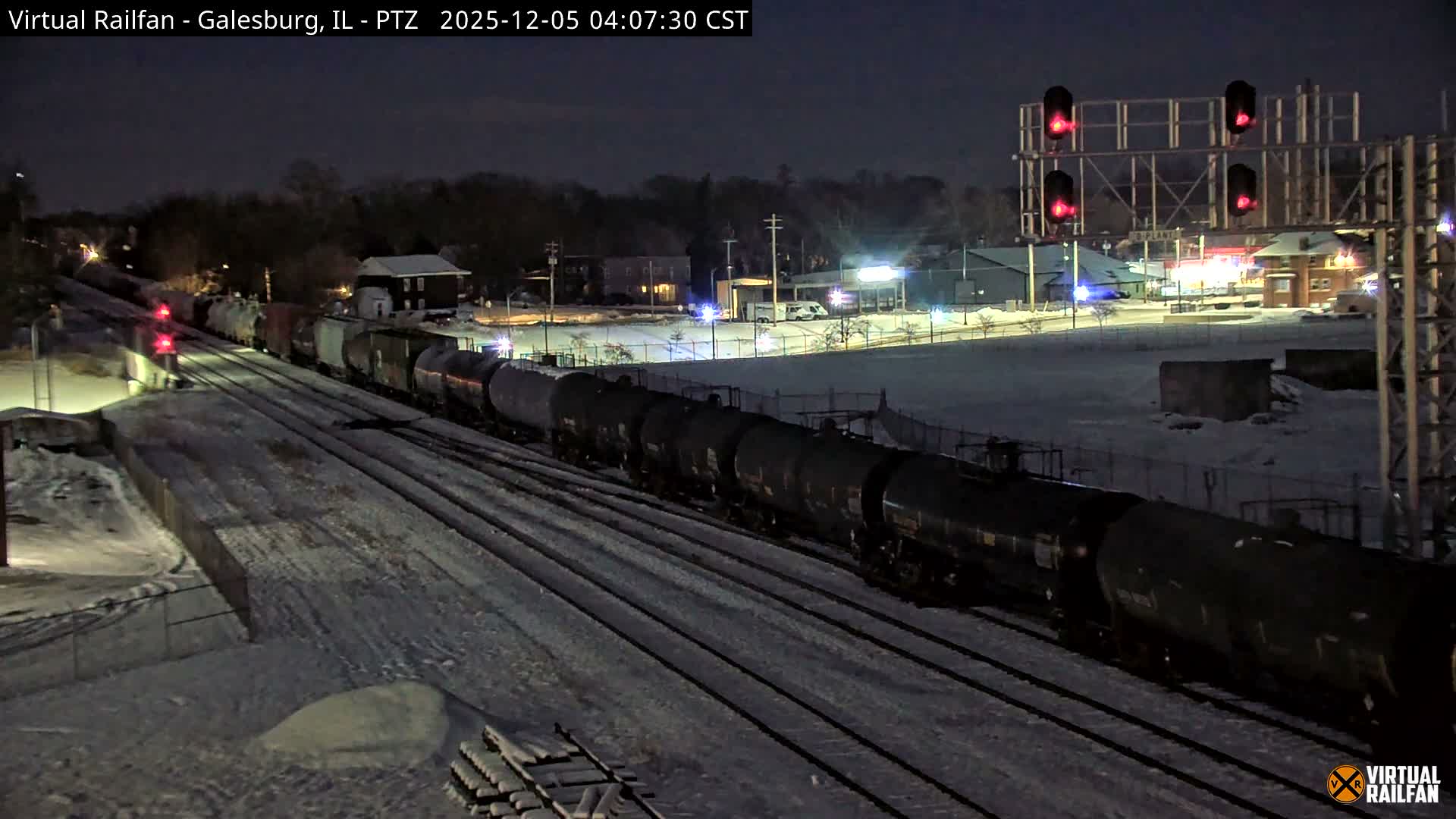 A long freight train, mostly consisting of dark tank cars, stretches along multiple snow-covered tracks at night, illuminated by streetlights and several bright red rail signals, under cold and snowy conditions.