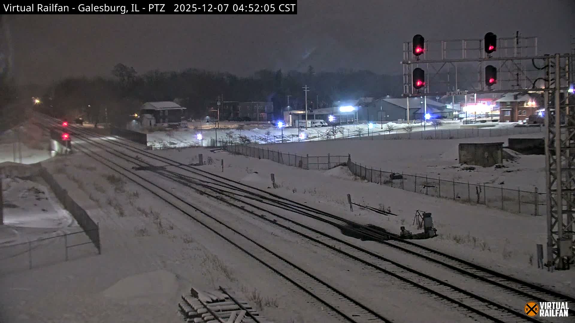 Multiple train tracks covered in snow stretch across a wintry night scene, illuminated by red signal lights and distant building lights under a dark sky.