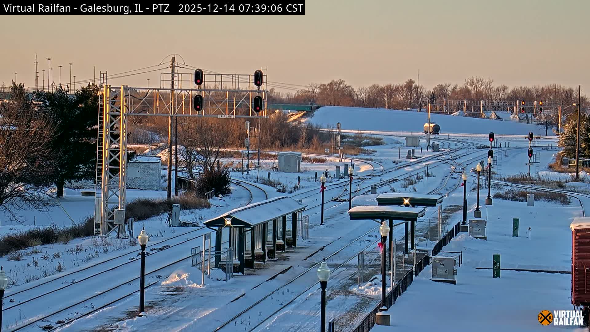 Multiple train tracks covered in snow stretch across a wintry night scene, illuminated by red signal lights and distant building lights under a dark sky.
