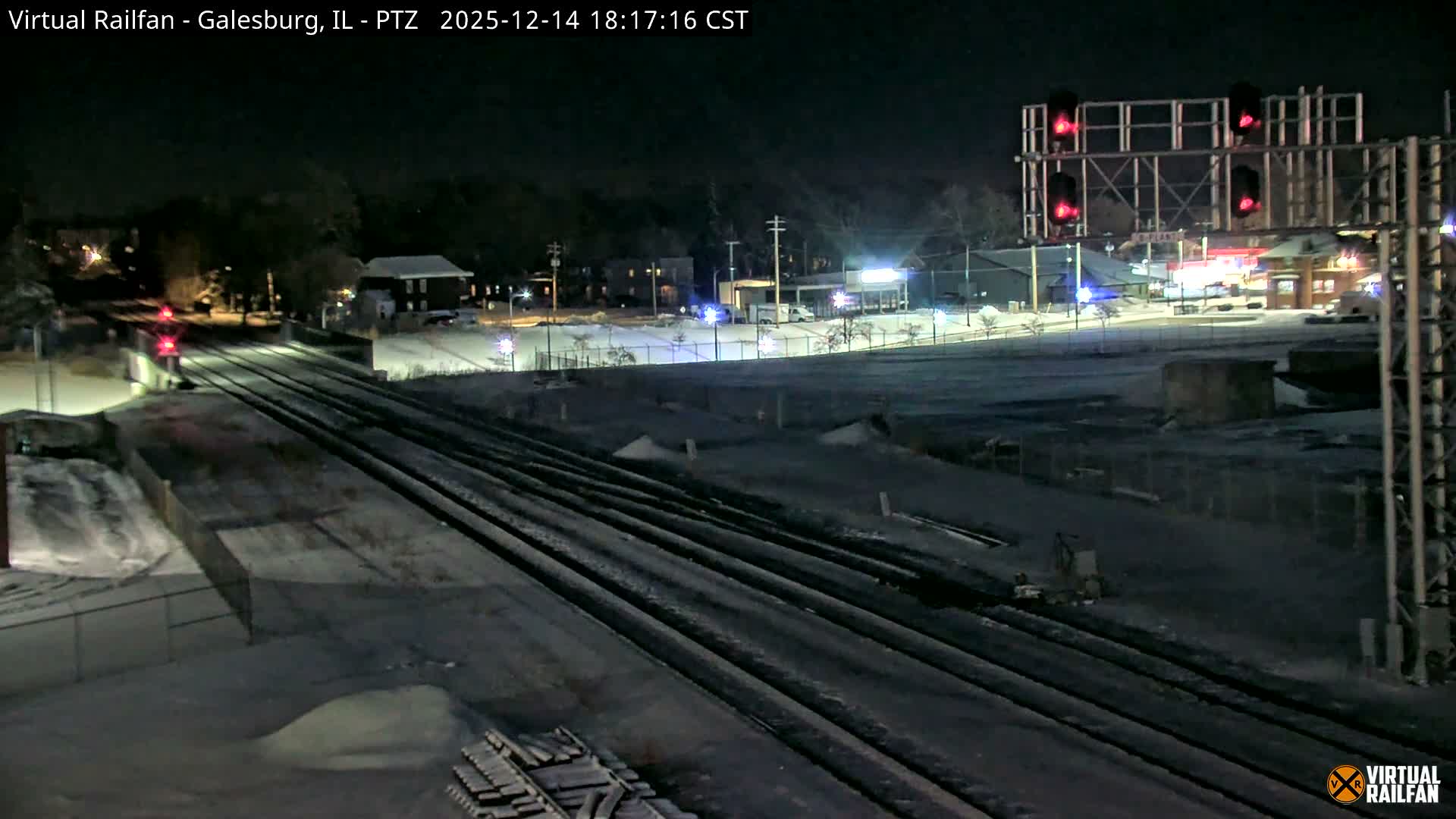 Multiple train tracks covered in snow stretch across a wintry night scene, illuminated by red signal lights and distant building lights under a dark sky.