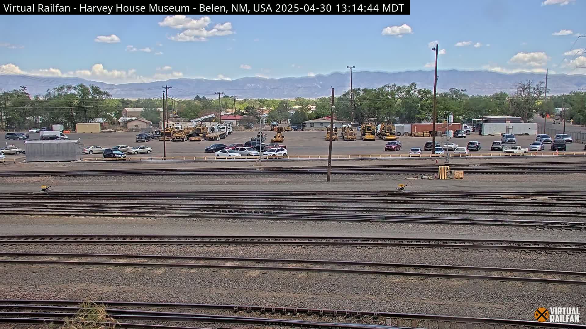 A sunny day reveals numerous parked cars and construction equipment near a set of parallel train tracks, with mountains visible in the background.