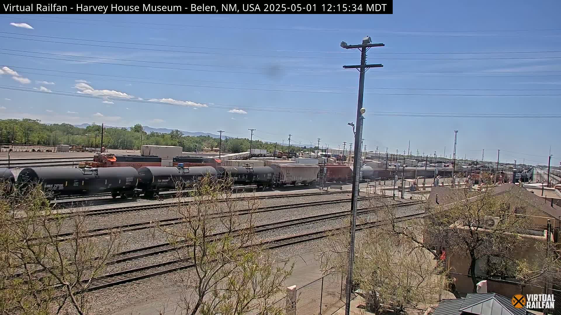 Under a mostly clear sky, a long freight train sits on a railway track next to a building, with mountains visible in the distance.
